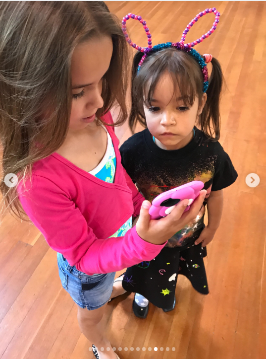 Mabel shows Fiona something on a pink phone as the sisters stand together indoors, with Fiona wearing a colorful bunny-ear headband and looking intently at the screen. The simple moment captures the girls sharing an everyday childhood activity.