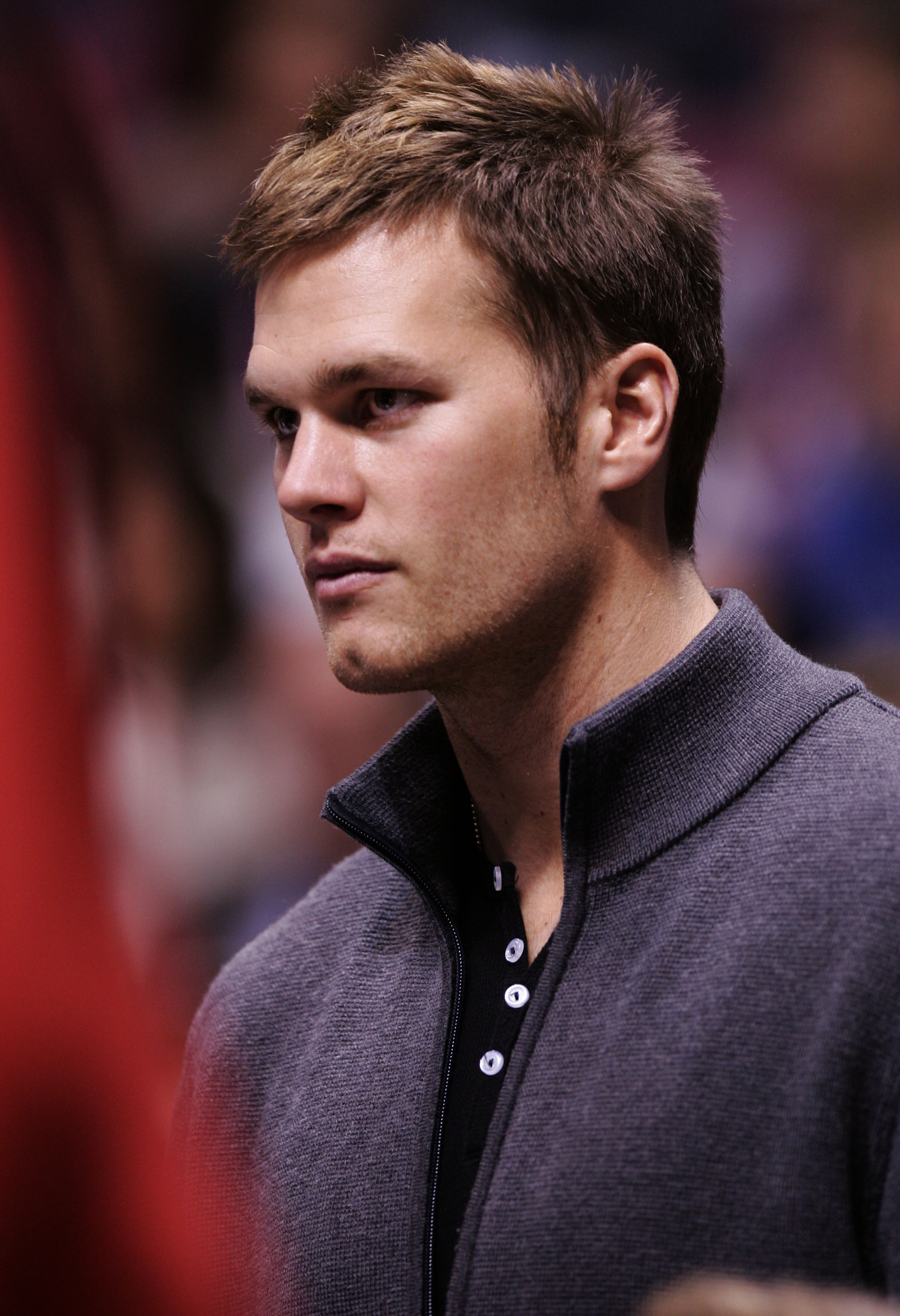 Tom Brady attends the Miami Heat vs. New Jersey Nets playoff game, sporting a black polo under a gray sweater. The shot highlights his chiseled jawline and effortlessly cool, spiked hair.