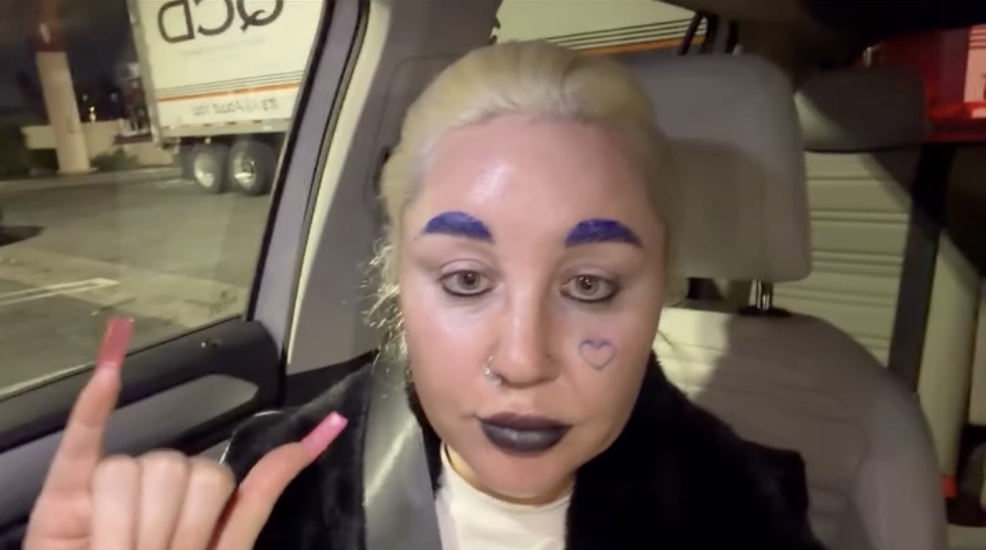 In January 2026, Amanda Bynes appears in a parked car with platinum blonde hair, purple brows, black lipstick, a heart on her cheek, and pink nails raised in a hand sign. | Source: Instagram/amandapandapandapanda1