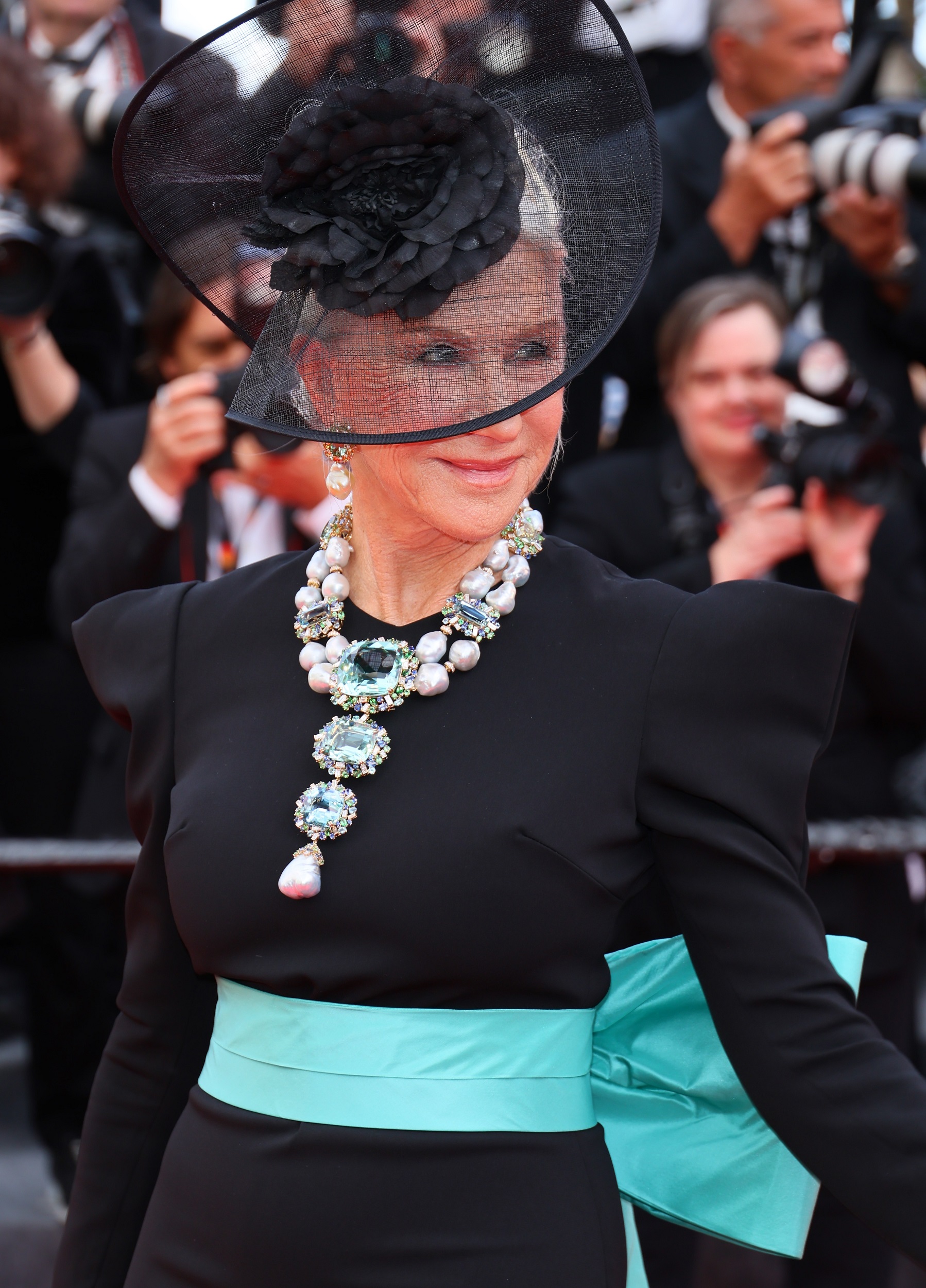 Mirren stuns in a structured black gown cinched with a bright aqua sash. Her statement necklace, anchored by a seafoam aquamarine gem, sparkles with sapphires, diamonds, green beryls, and bold baroque pearls, making a dramatic and elegant impression.
