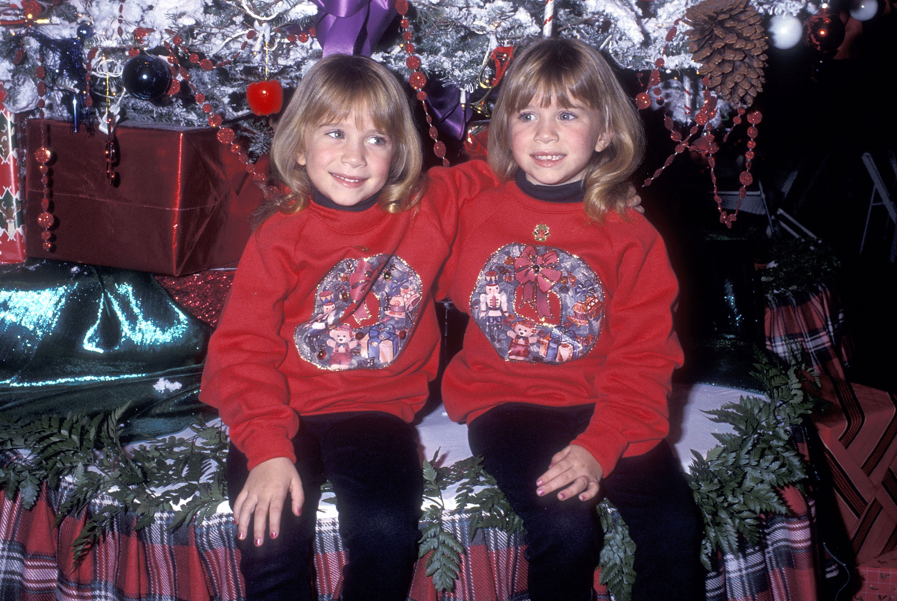 Cheerful, wider grins and matching rounded blonde bobs highlighted the sisters at 6 years old during the holiday parade on November 29, 1992, where their festive bright red sweatshirts with embroidered wreaths and teddy bears layered over black turtlenecks signaled an increasingly animated stage presence.
