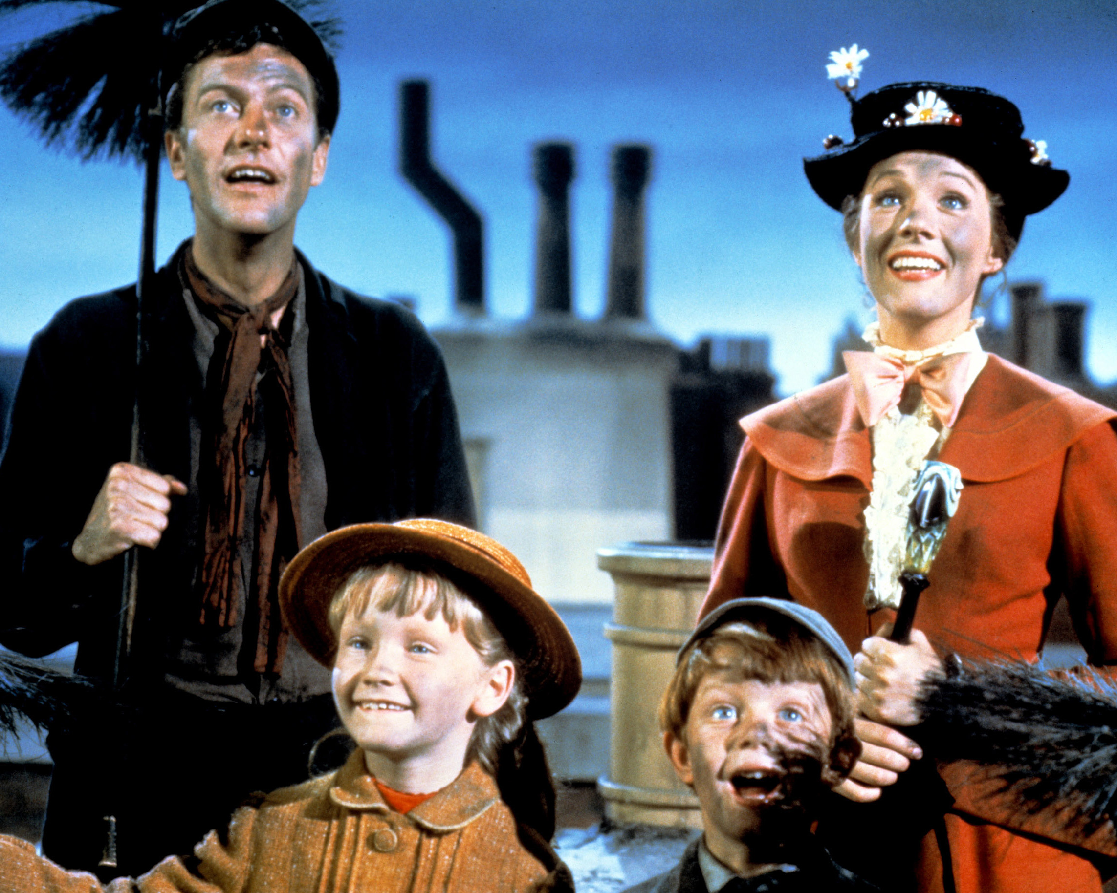 Julie Andrews leads a whimsical rooftop scene as the magical nanny in Walt Disney's 1964 classic musical "Mary Poppins," smiling with soot-dusted cheeks alongside co-star Dick Van Dyke as chimney sweep Bert, Karen Dotrice as Jane Banks, and Matthew Garber as Michael Banks. Wearing her flower-trimmed black boater hat, burnt-orange coat with a ruffled collar, and clutching her iconic parrot-head umbrella, Andrews looks skyward alongside her chimney-sweeping companions high above London's rooftops.