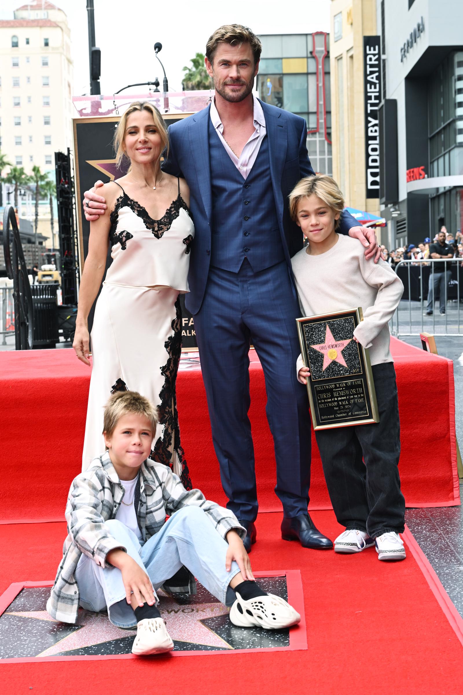 Chris Hemsworth poses with his wife, Elsa Pataky, and their twin sons, Tristan and Sasha, during his Hollywood Walk of Fame ceremony on May 23, 2024, in Los Angeles. Elsa wore a cream satin dress with black lace accents, while Chris appeared in a sharp blue suit. Tristan held the commemorative plaque, and Sasha sat near his father's newly unveiled star, dressed casually in light denim and a plaid shirt.