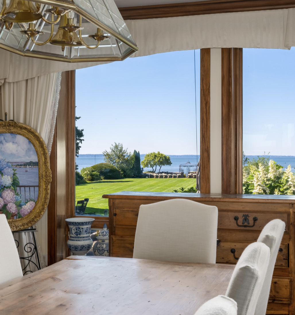 The dining area features a rustic wooden table, upholstered chairs, and large windows overlooking the manicured lawn and Long Island Sound — a peaceful setting for family meals with a spectacular waterfront view. | Source: Instagram/@brizolisjanzen