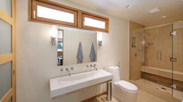 A modern bathroom with a walk-in shower. A long white basin sits beneath a large mirror, while a glass shower enclosure with beige stone tiles occupies the right side of the room. Chrome fixtures and wooden trim complement the neutral interior. | Source: Redfin