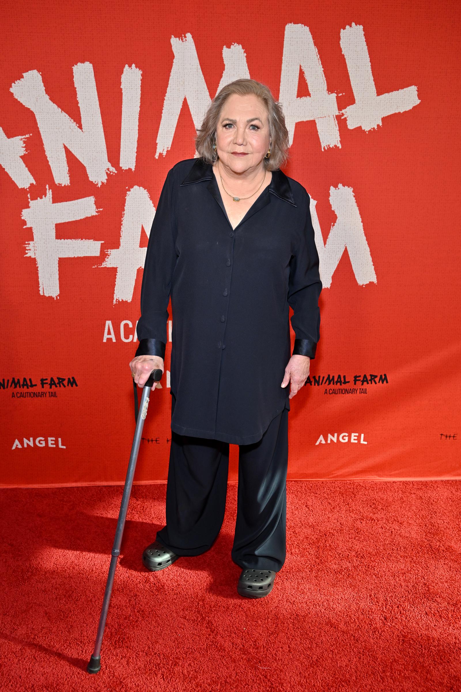 Kathleen Turner poses for a full-length photo at the "Animal Farm" premiere, dressed in a monochromatic navy outfit and dark slip-on shoes. She holds a cane while standing in front of the film's red promotional backdrop.