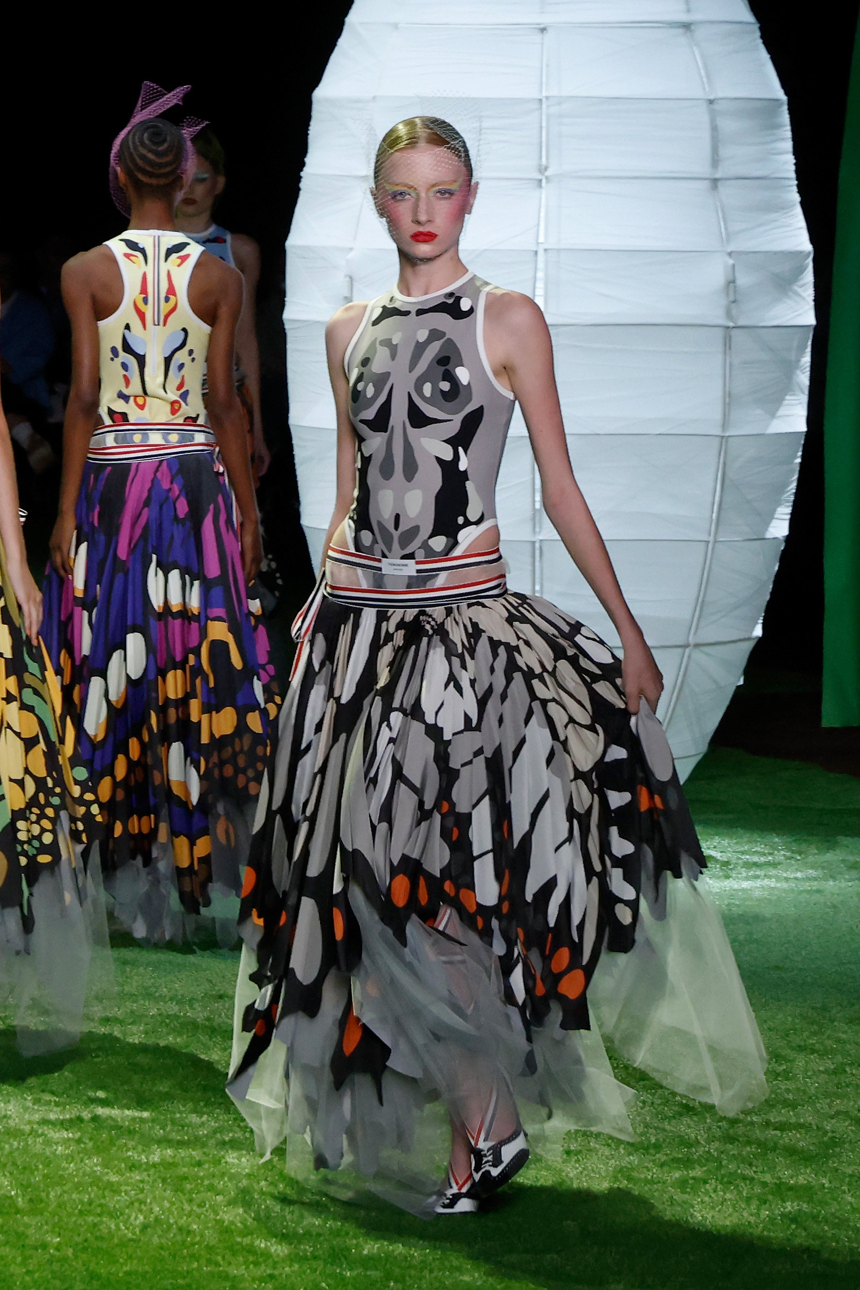 Kai Schreiber strutted forward in full motion along the artificial lawn runway during the Thom Browne presentation for New York Fashion Week on September 15, 2026, in New York City. The emerging runway talent captured the spotlight wearing a monochrome patterned tank, red-striped trim, and a cascading butterfly-motif midi skirt layered with delicate mesh over two-tone oxford heels. | Source: Getty Images