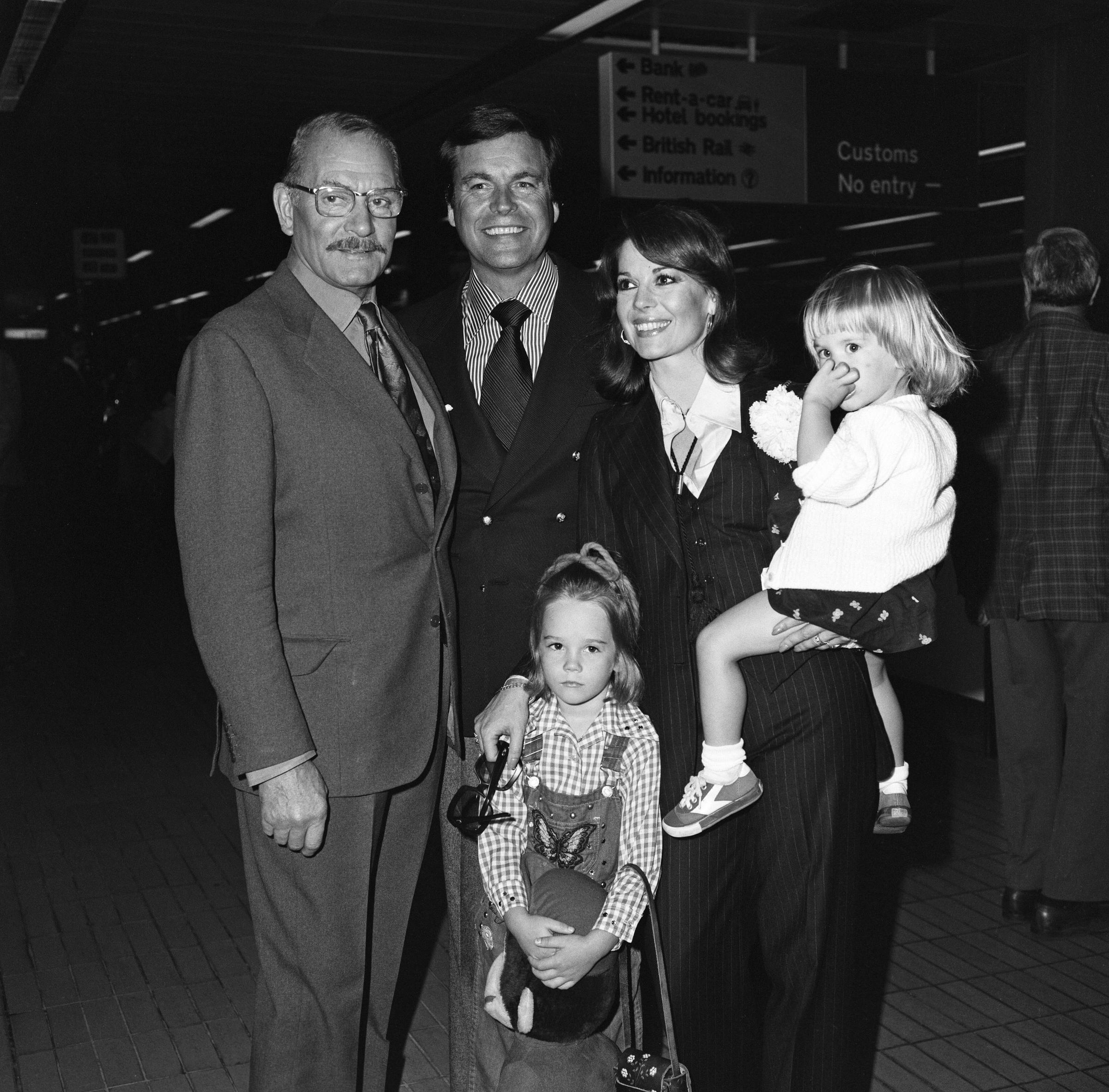 Robert Wagner and Natalie Wood arrive at Heathrow Airport on May 19, 1976, with daughters Natasha and Courtney. Natalie carries Courtney, who clutches a doll, while Natasha stands in front holding a stuffed toy. The family poses with Laurence Olivier, ahead of filming a Granada TV production of "Cat on a Hot Tin Roof."