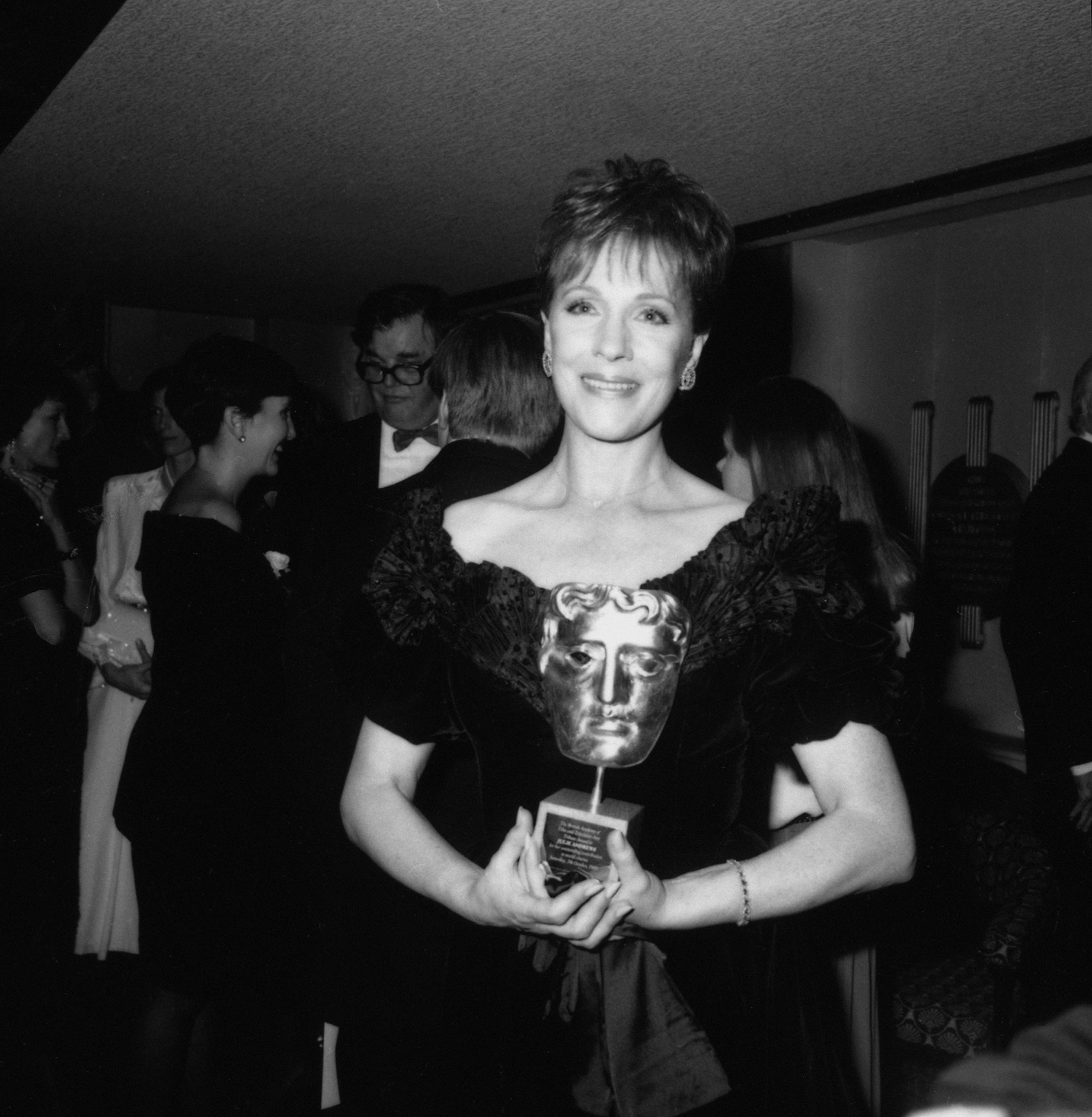 Julie Andrews proudly holds her BAFTA award during a special tribute evening celebrating her career, held at the Odeon in Leicester Square, London, on October 7, 1989. The actress glows in an off-the-shoulder black velvet gown featuring a dramatic ruffled neckline, accessorized with diamond drop earrings and a delicate bracelet.