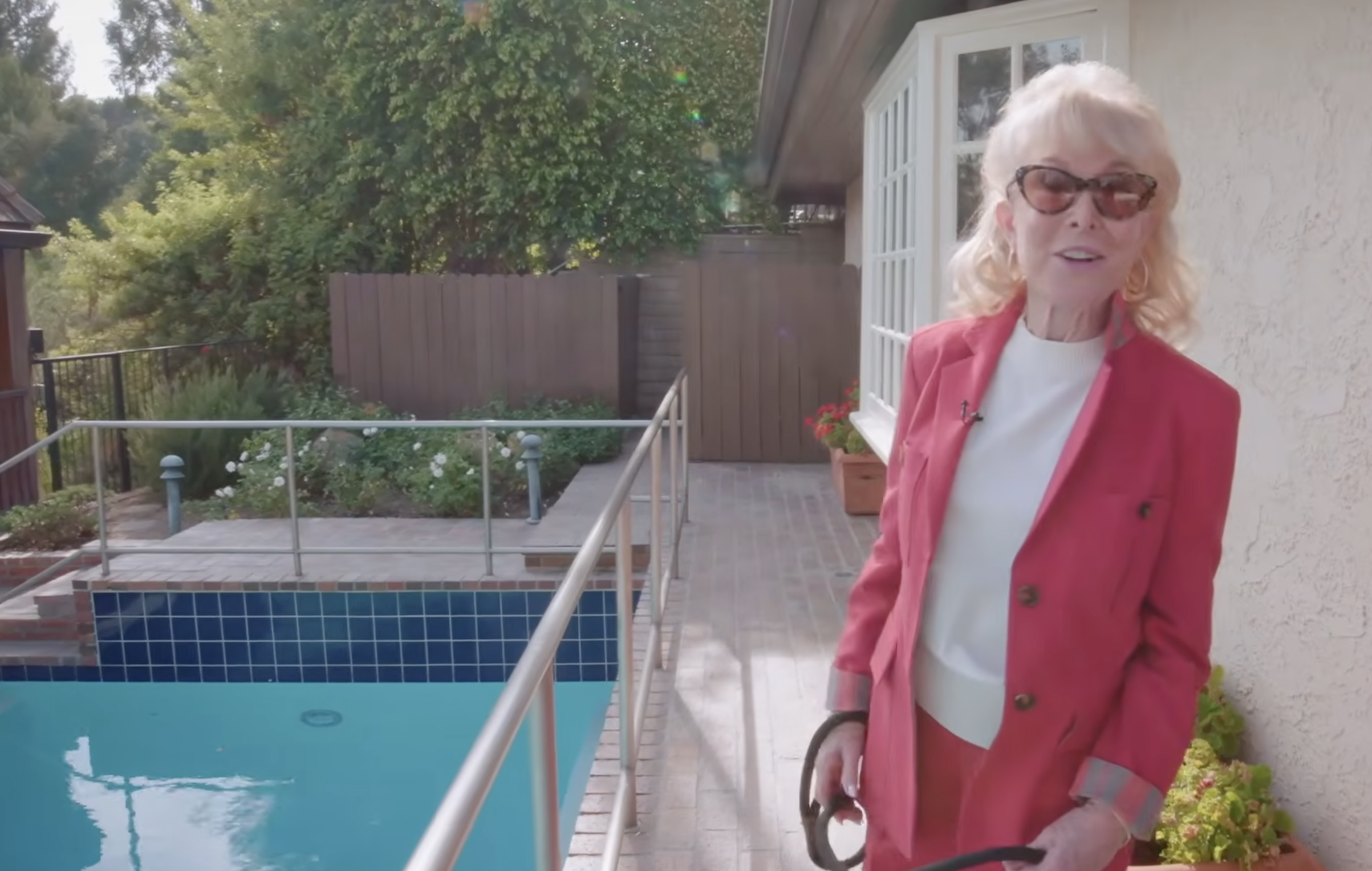 Barbara Eden stands beside her swimming pool wearing sunglasses and a pink-red pantsuit, holding a garden hose as she smiles toward the camera. The turquoise water, blue pool tiles, metal safety railing, and surrounding greenery highlight the home's outdoor space. | Source: YouTube/People