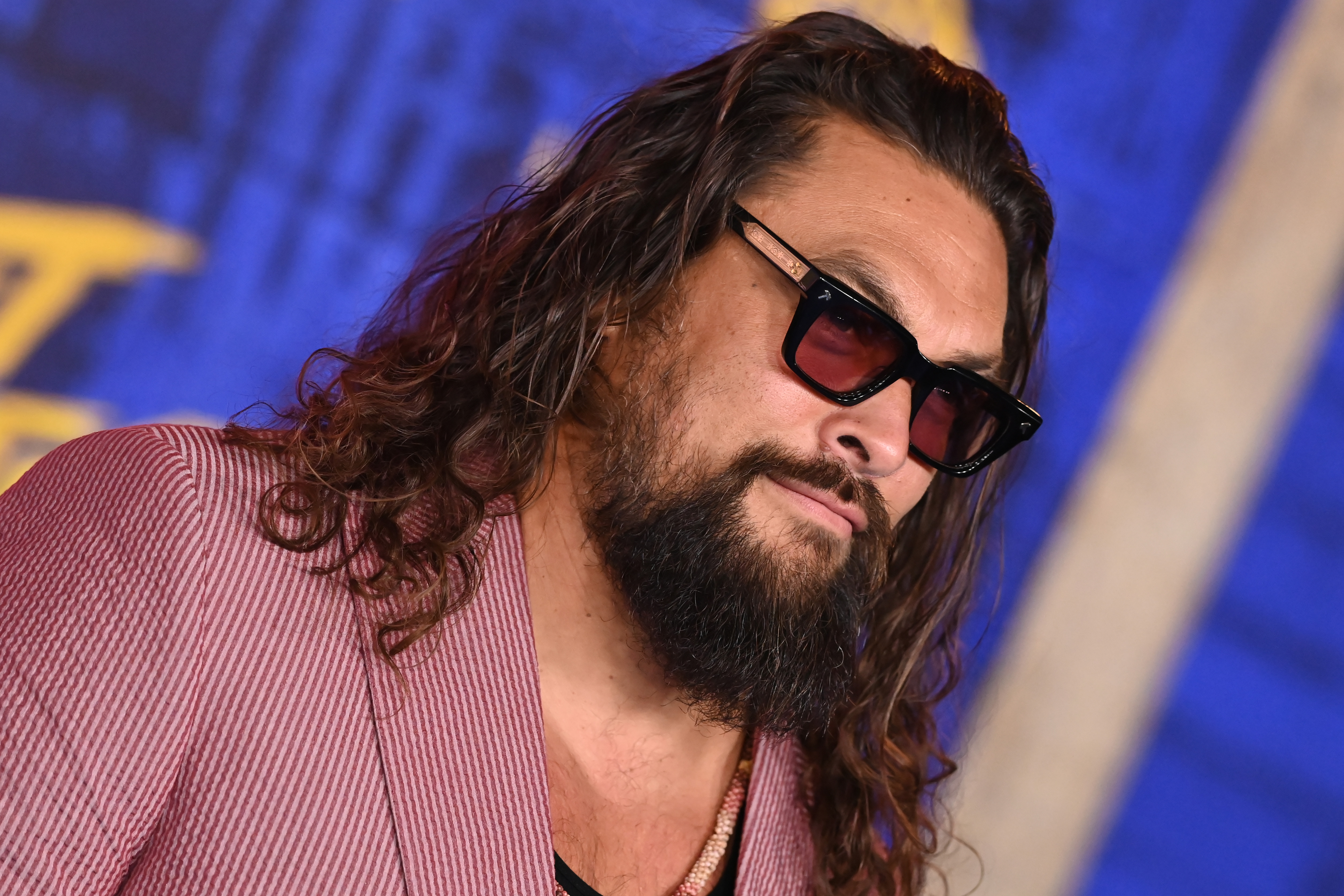 Jason Momoa leans slightly in a close-up portrait at the Los Angeles premiere of Paramount Pictures' "Street Fighter" at the TCL Chinese Theatre on October 12, 2026, in Hollywood, California. His wavy brown mane tumbles over his shoulder, highlighting tinted rectangular sunglasses, a full beard, and a delicate beaded necklace resting beneath a textured dusty-rose notch-lapel jacket. | Source: Getty Images