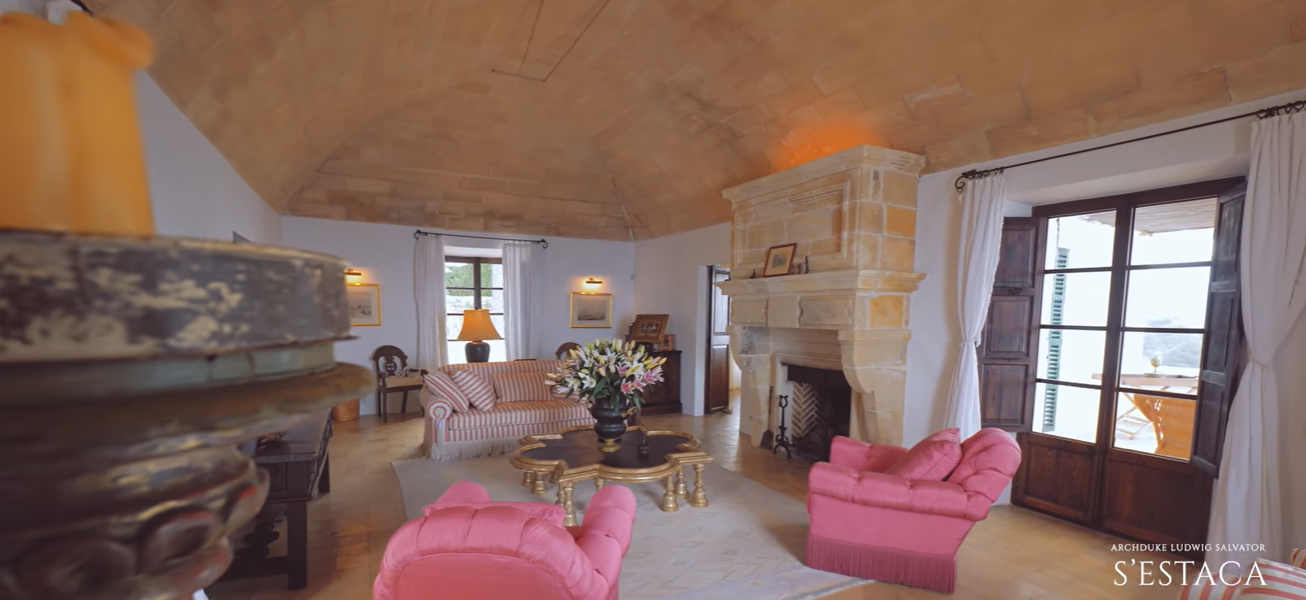 A high stone ceiling and large fireplace bring old-world character to this sitting room. Pink chairs and a striped sofa add color, while glass doors open the space toward an outdoor terrace. | Source: YouTube/billionsluxuryportal