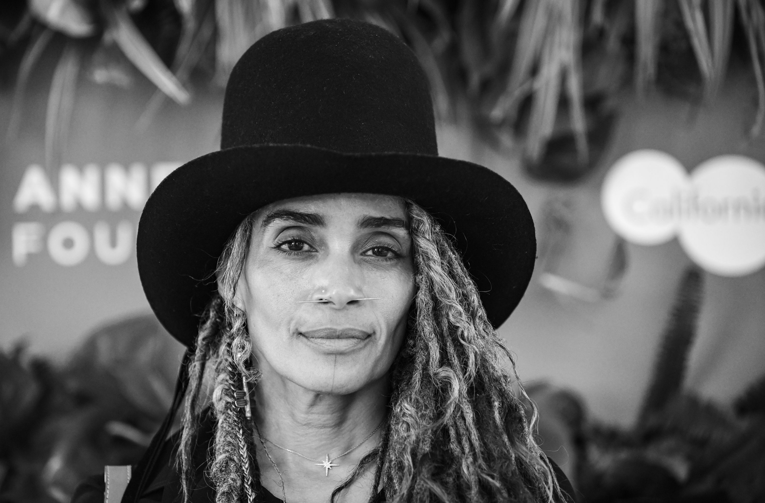 Lisa Bonet poses for a close-up portrait while attending a VIP premiere event in Los Angeles, wearing a tall black hat and long textured locs that frame her face.