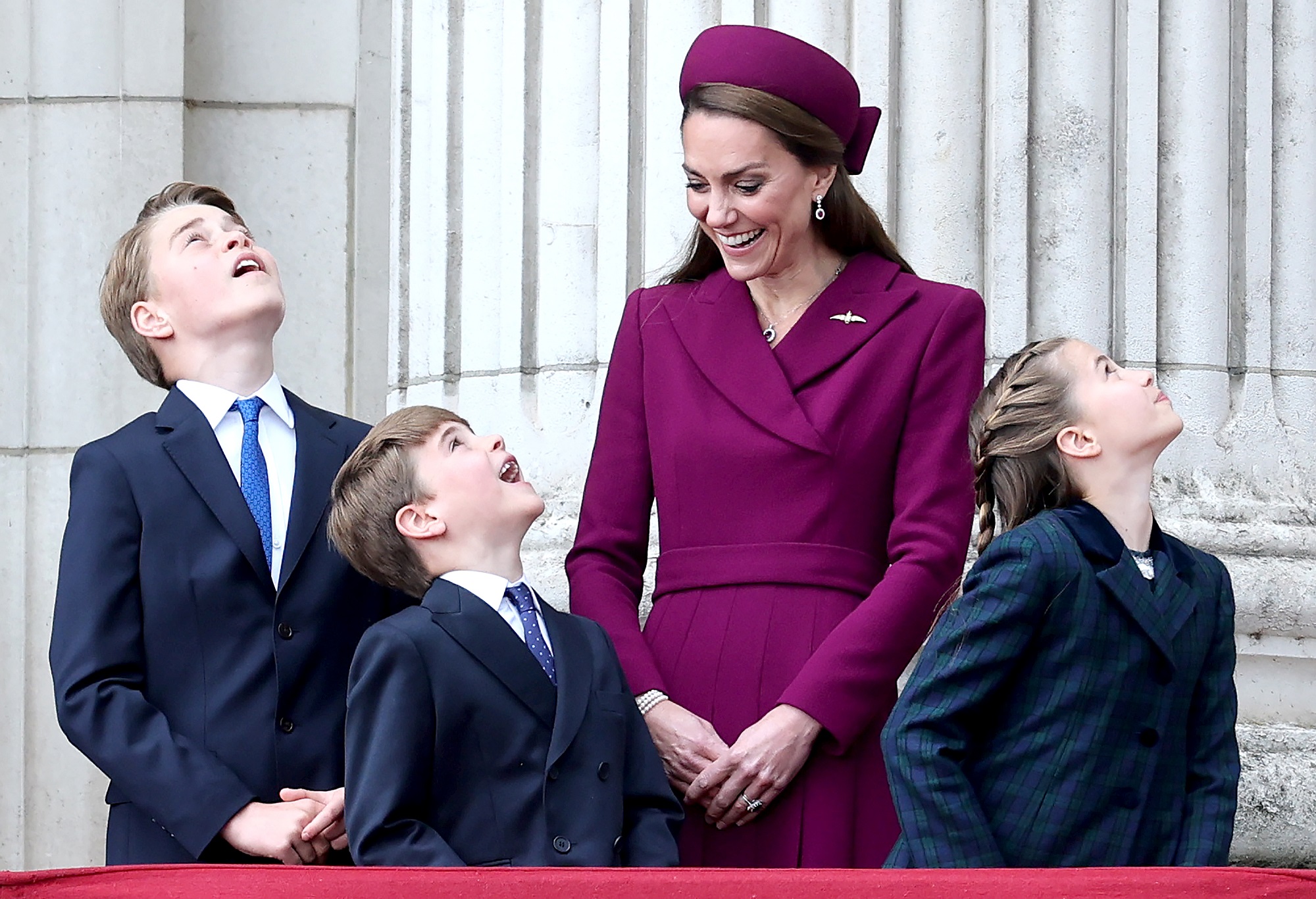Standing on the balcony, Charlotte, George, and Louis gaze upward in unison, likely watching a flypast, their faces lit with wonder and curiosity. Beside them, Catherine beams with joy, clearly amused by their expressions and sharing in the moment of excitement.
