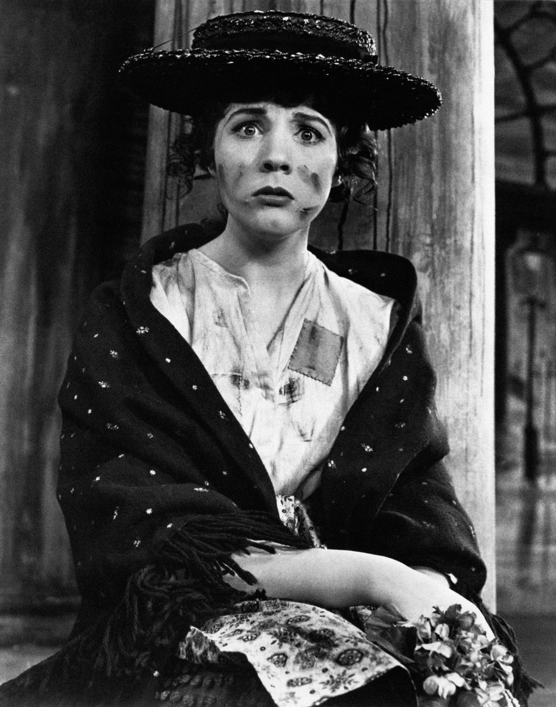 Julie Andrews captures the gritty charm of Eliza Doolittle in a scene from the stage musical "My Fair Lady," sitting with a wide-eyed, sullen expression and soot smudges across her cheeks while dressed in a straw boater hat, a fringed dark shawl draped over a patched blouse, and holding a small bunch of violets.