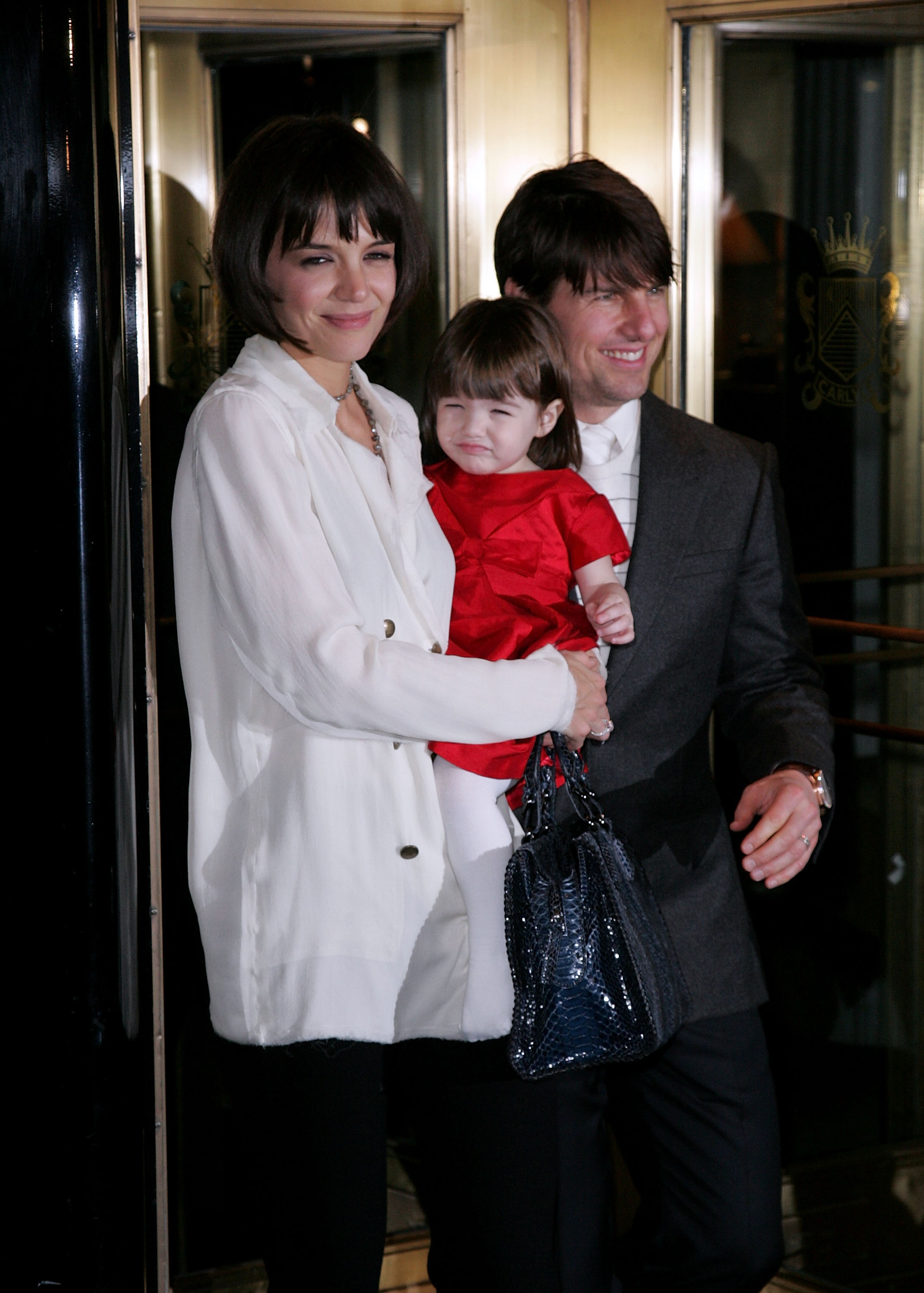 In a sweet family outing, Katie Holmes cradled a giggling Suri Cruise, who looked festive in a red dress, while Tom Cruise stood beside them beaming with pride. Katie, chic in a white coat and bob hairstyle, exuded modern elegance and maternal warmth.