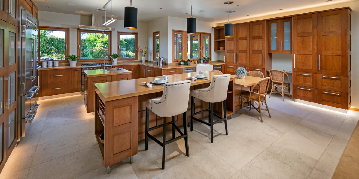 The large wooden kitchen. Extensive wooden cabinets line the walls beneath warm lighting, while stone countertops and a central island provide ample workspace. A long counter with upholstered bar stools adds to the kitchen's welcoming, functional design. | Source: Redfin