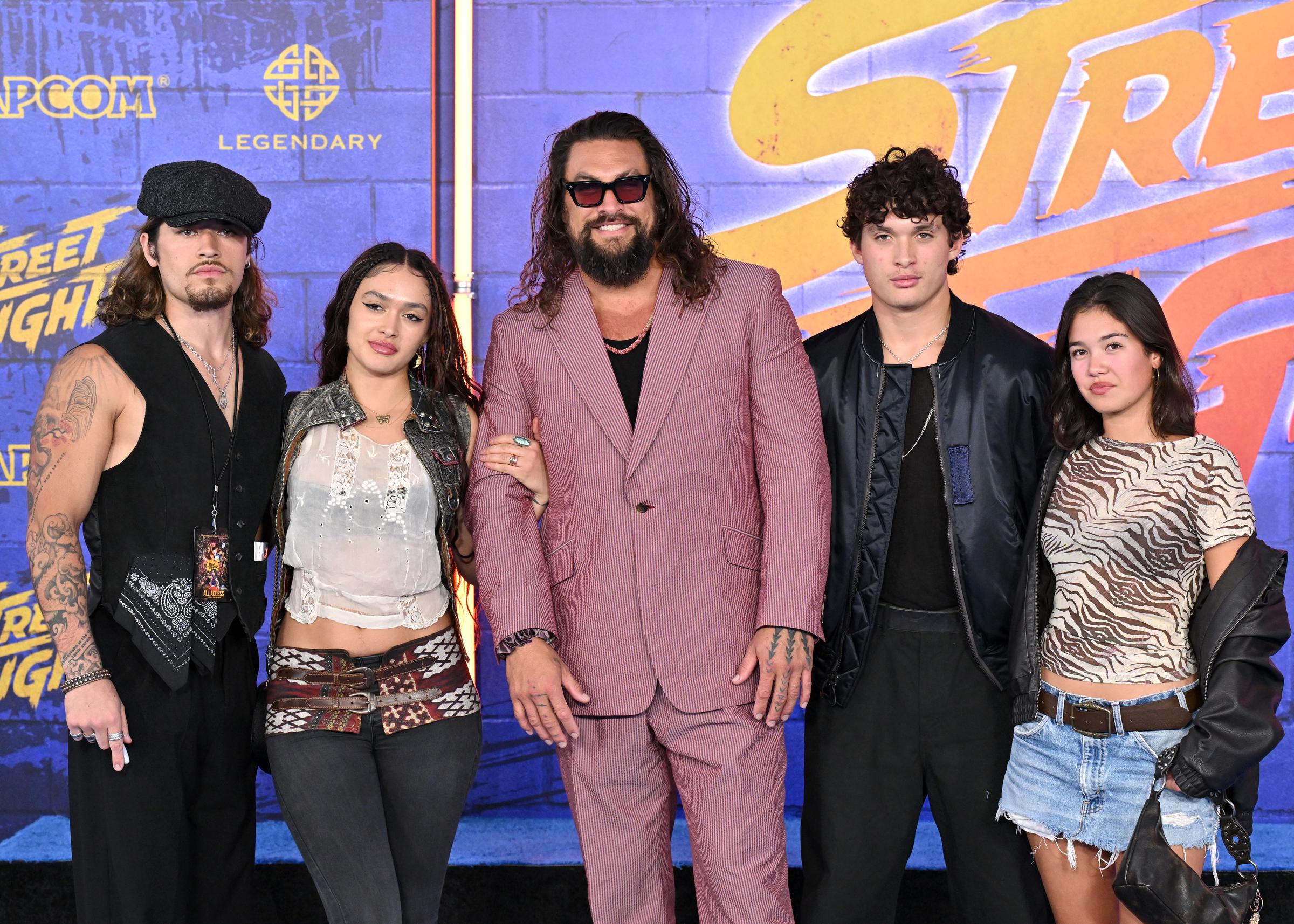 Positioned squarely in front of the Capcom and Legendary production logos on the press wall, the entire group lines up facing forward in a clean, balanced composition. Nui Momoa anchors the left side in an all-black ensemble featuring a newsboy cap, vest, and layered necklaces, balancing the casual edge of the denim shorts, tiger-stripe tee, and draped leather jacket on the far right. | Source: Getty Images