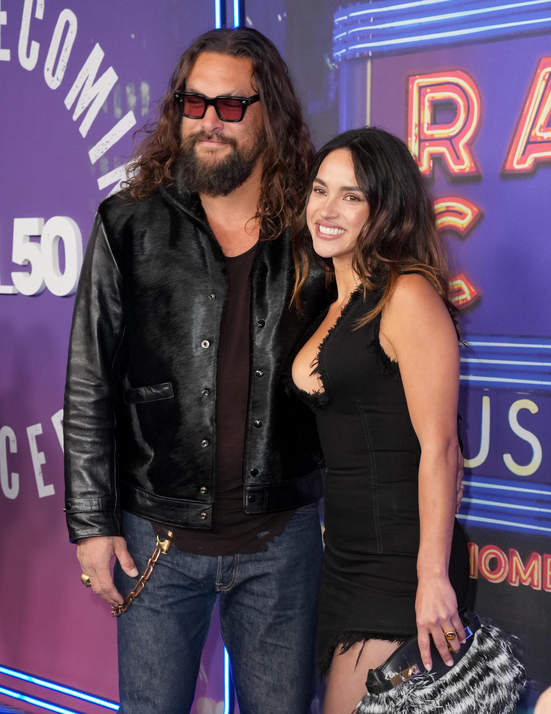 Jason Momoa and Adria Arjona smile side by side at SNL50: The Homecoming Concert at Radio City Music Hall on February 14, 2025, in New York City. Momoa wears dark jeans with a chunky brass pocket chain, a distressed brown undershirt, a sleek glossy black jacket, and red-tinted sunglasses, posing close to Arjona, who wears a sleeveless black mini dress with raw fringed trim and carries a furry textured clutch purse. | Source: Getty Images