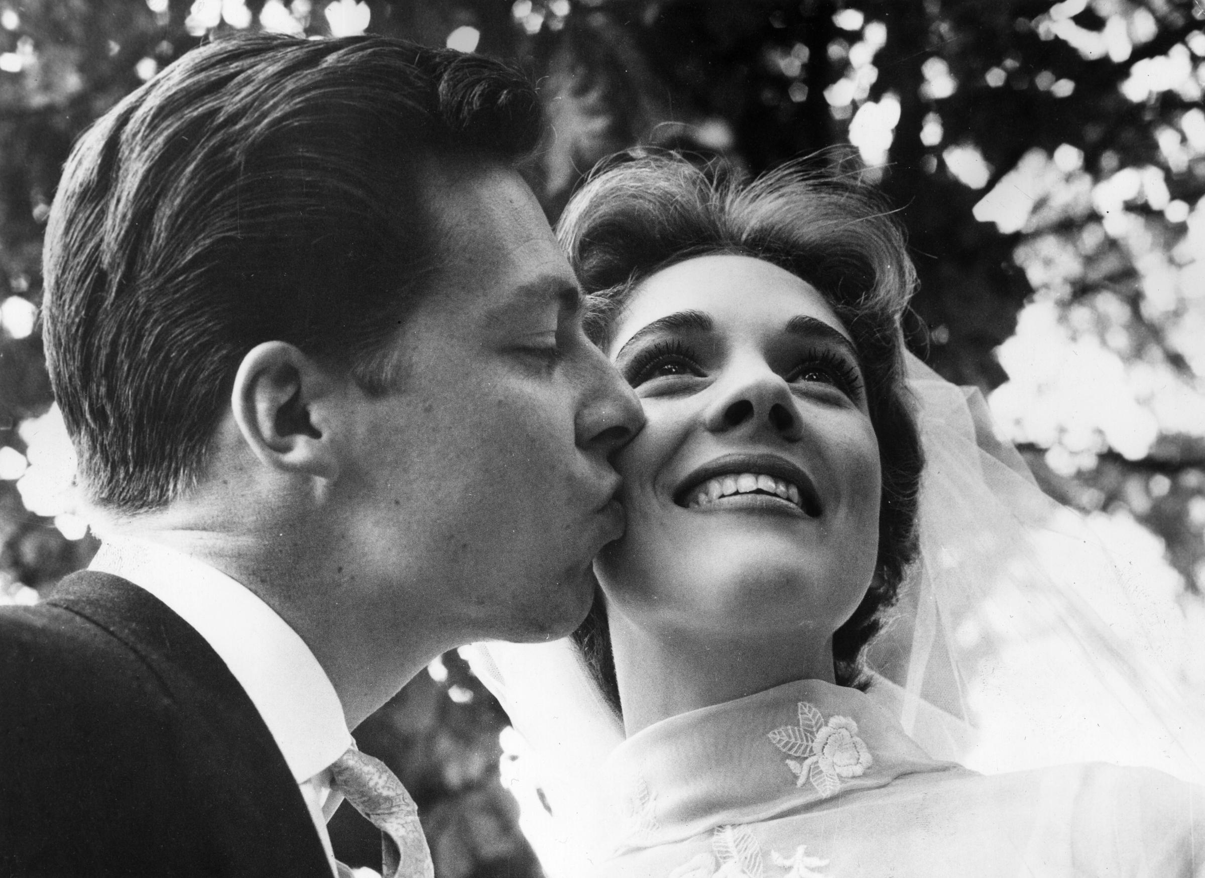 British costume designer Tony Walton kisses the cheek of his radiant new bride, Julie Andrews, on their wedding day in England on May 10, 1959. The smiling actress and singer glows with joy, wearing an elegant high-collared bridal gown accented with delicate floral embroidery and a sheer tulle veil draped over her styled brunette curls.