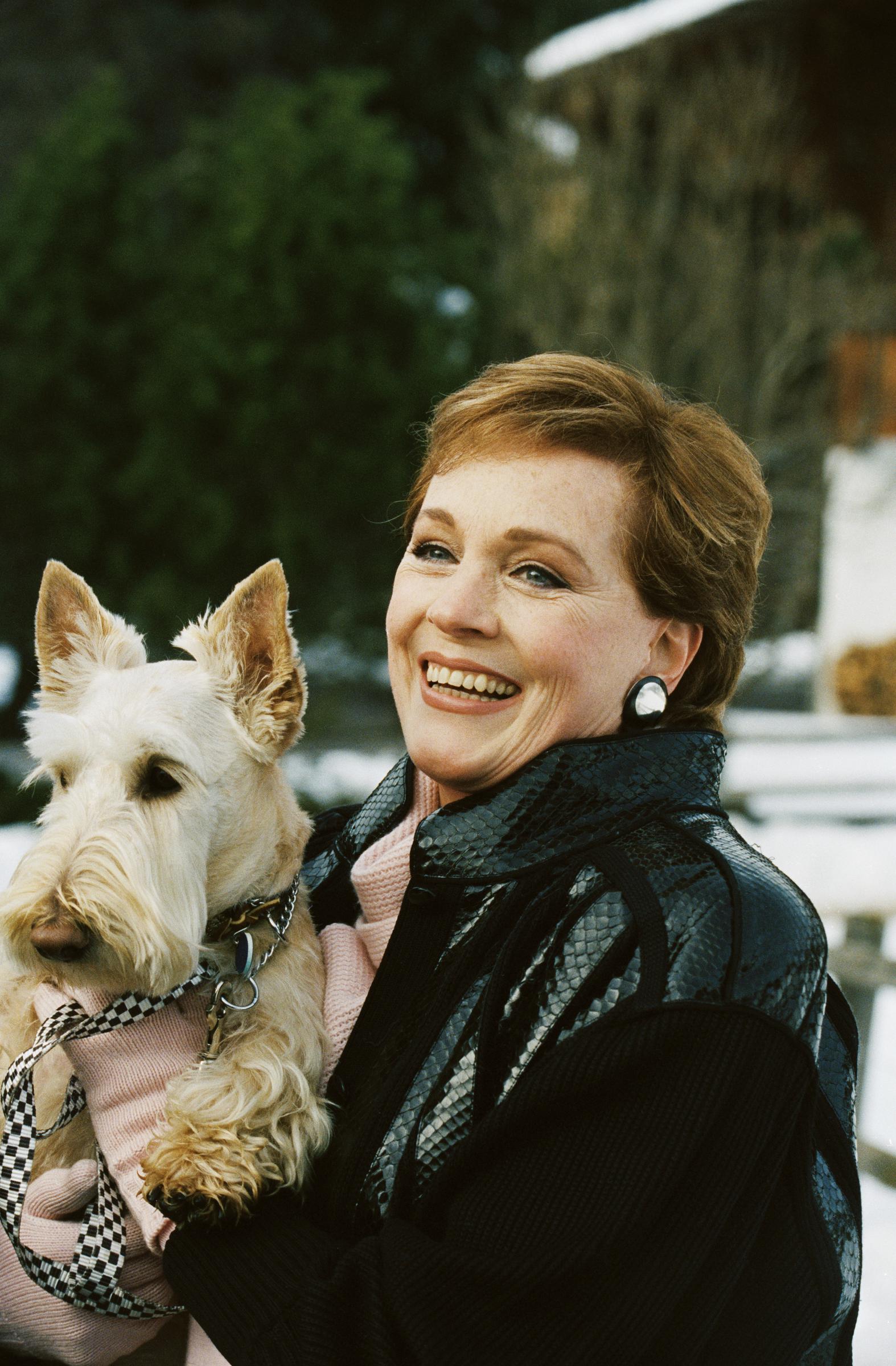 Julie Andrews shares a joyful outdoor moment holding her light-colored Scottish terrier in a portrait dated December 31, 1993. Set against a winter backdrop with dusted snow, the smiling actress stays warm in a pink knit sweater paired with a chic black snakeskin-textured jacket, finished with round statement stud earrings.