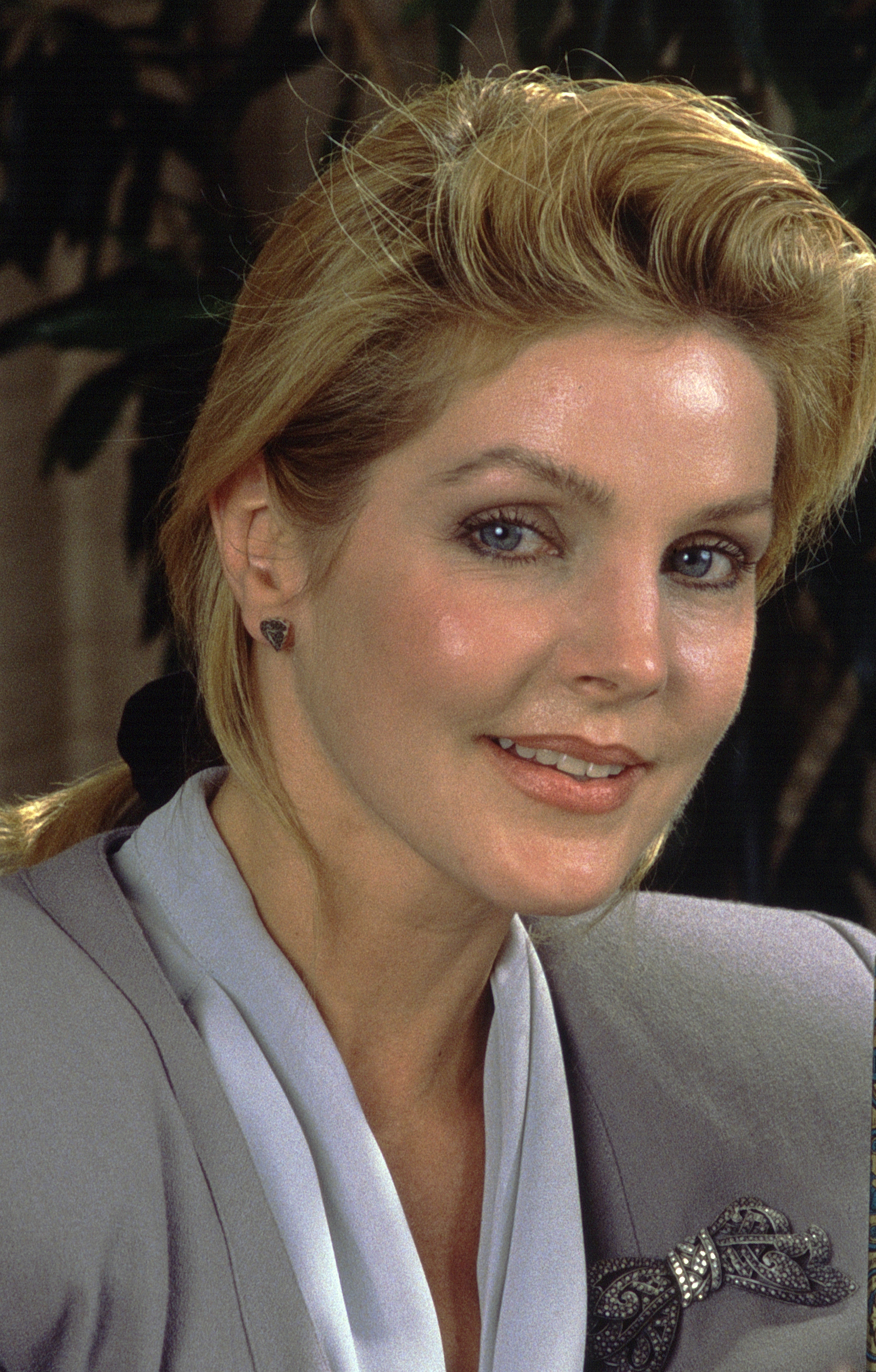 Priscilla Presley poses for a portrait during the unveiling of a new fragrance in Los Angeles, California, on May 13, 1988, dressed in a tailored gray blazer accented with an ornate jeweled bow brooch over a soft white draped blouse, with her blonde-highlighted hair swept back into a low ponytail secured with a dark scrunchie.