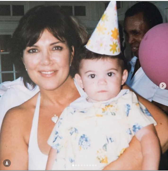 A young Kris Jenner holds baby Kylie Jenner during a birthday celebration, with Kylie wearing a floral outfit and a festive party hat. Kris smiles at the camera while cradling her youngest daughter in her arms. | Source: Instagram/krisjenner
