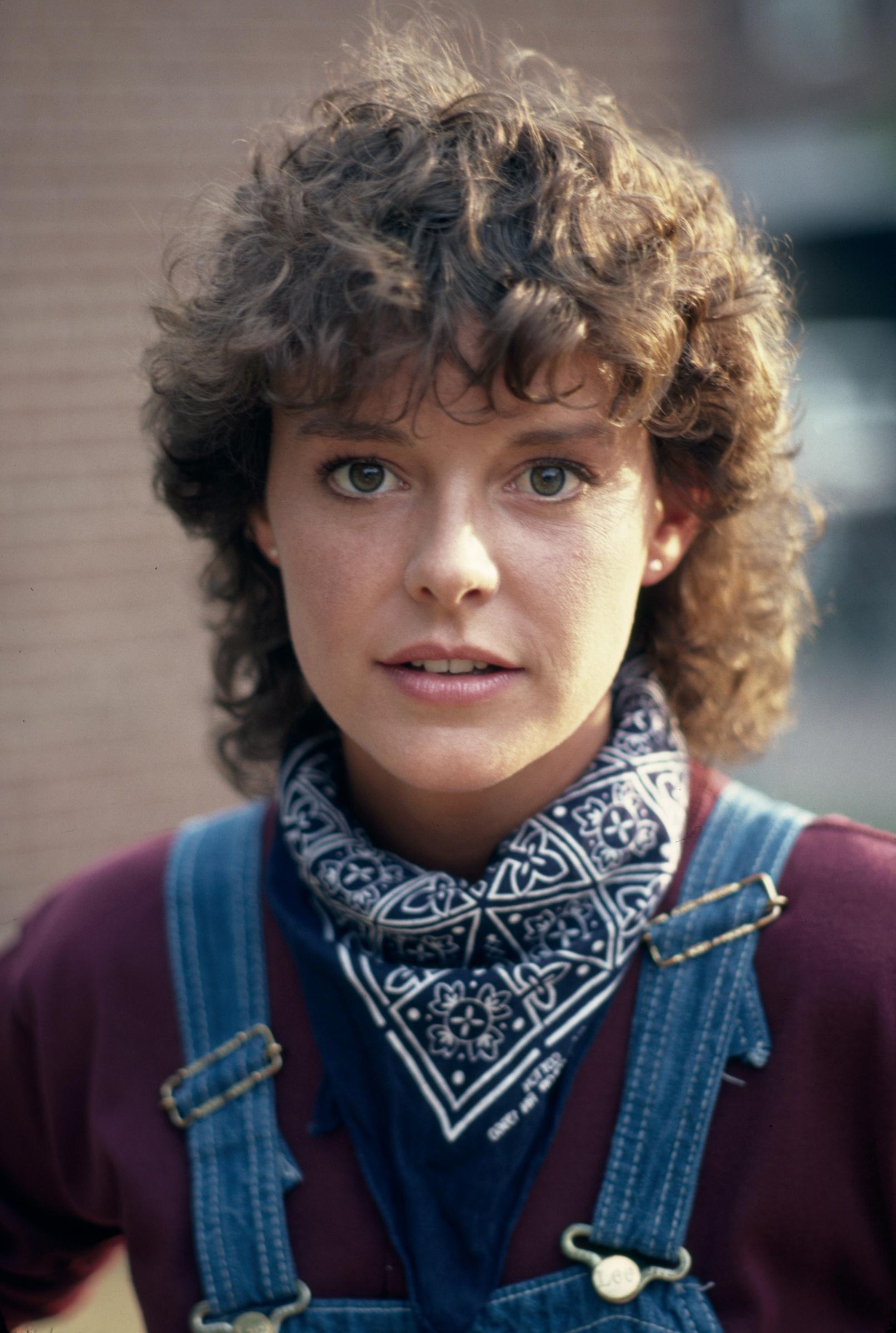 Amanda Bearse posed in character as Karen for the television movie "First Affair" on October 25, 1983, while filming on location in Boston, Massachusetts. Sporting voluminous, curly brunette locks with cropped bangs, the actress embraced an early '80s collegiate style in classic denim bib overalls over a maroon long-sleeve top, tied off with a patterned blue-and-white bandana around her neck. | Source: Getty Images