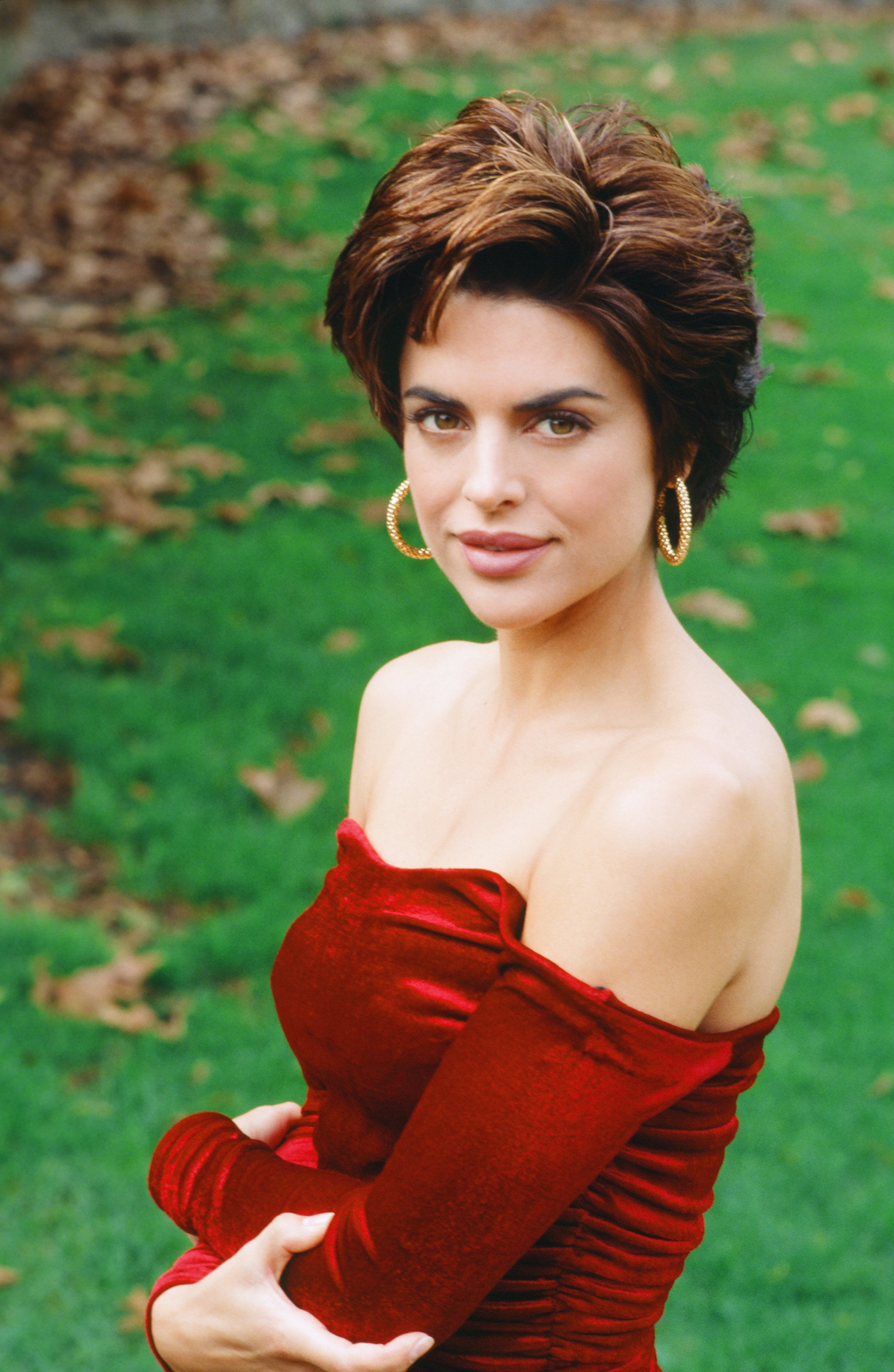 Lisa Rinna poses outdoors in a red off-the-shoulder velvet dress and gold hoop earrings. Her short, voluminous brunette hairstyle offers a sleeker variation of the layered cut that became her signature during the 1990s. | Source: Getty Images.