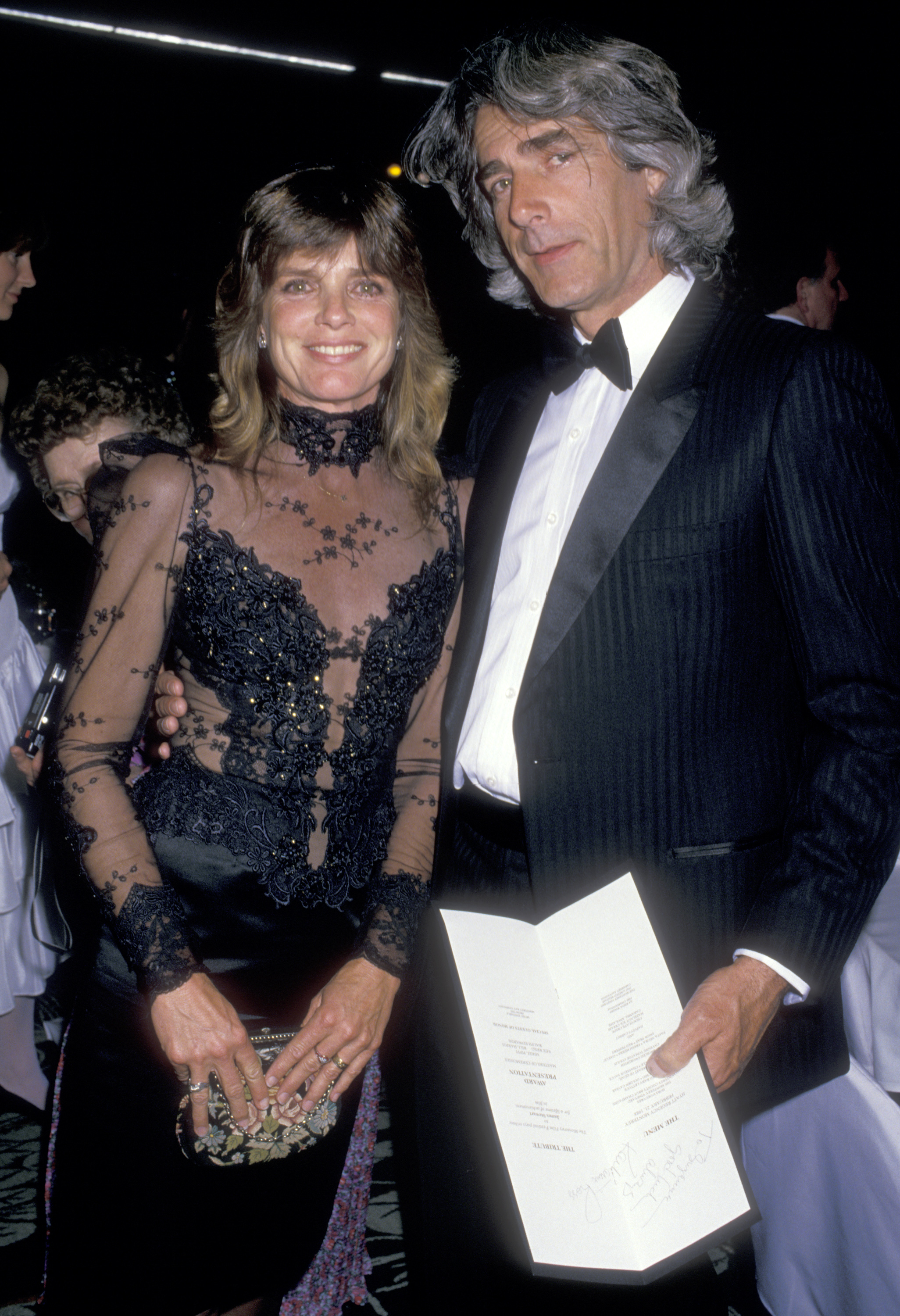 Katharine Ross and Sam Elliott looked elegant at the First Annual Golden Cypress Award Honoring James Stewart on February 21, 1988, in Monterey, California. Katharine wore a sheer black lace gown with intricate embroidery and a high collar, while Sam dressed in a pinstriped tuxedo with a black bow tie, holding an event program as they posed together. | Source: Getty Images