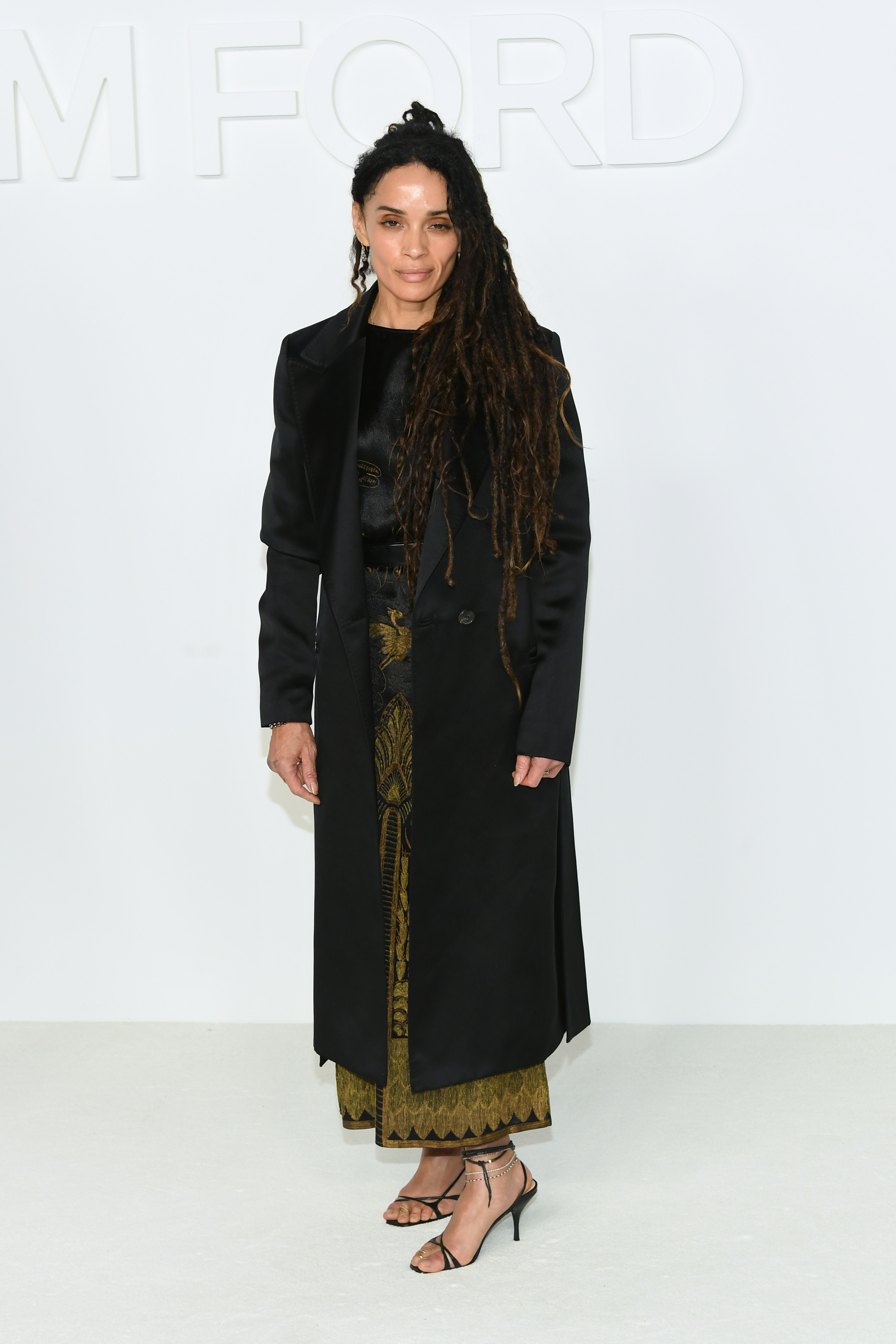 Lisa Bonet attends the Tom Ford AW20 show in Hollywood, pairing a long black coat with a patterned dress and strappy heels as she poses along the runway venue backdrop.