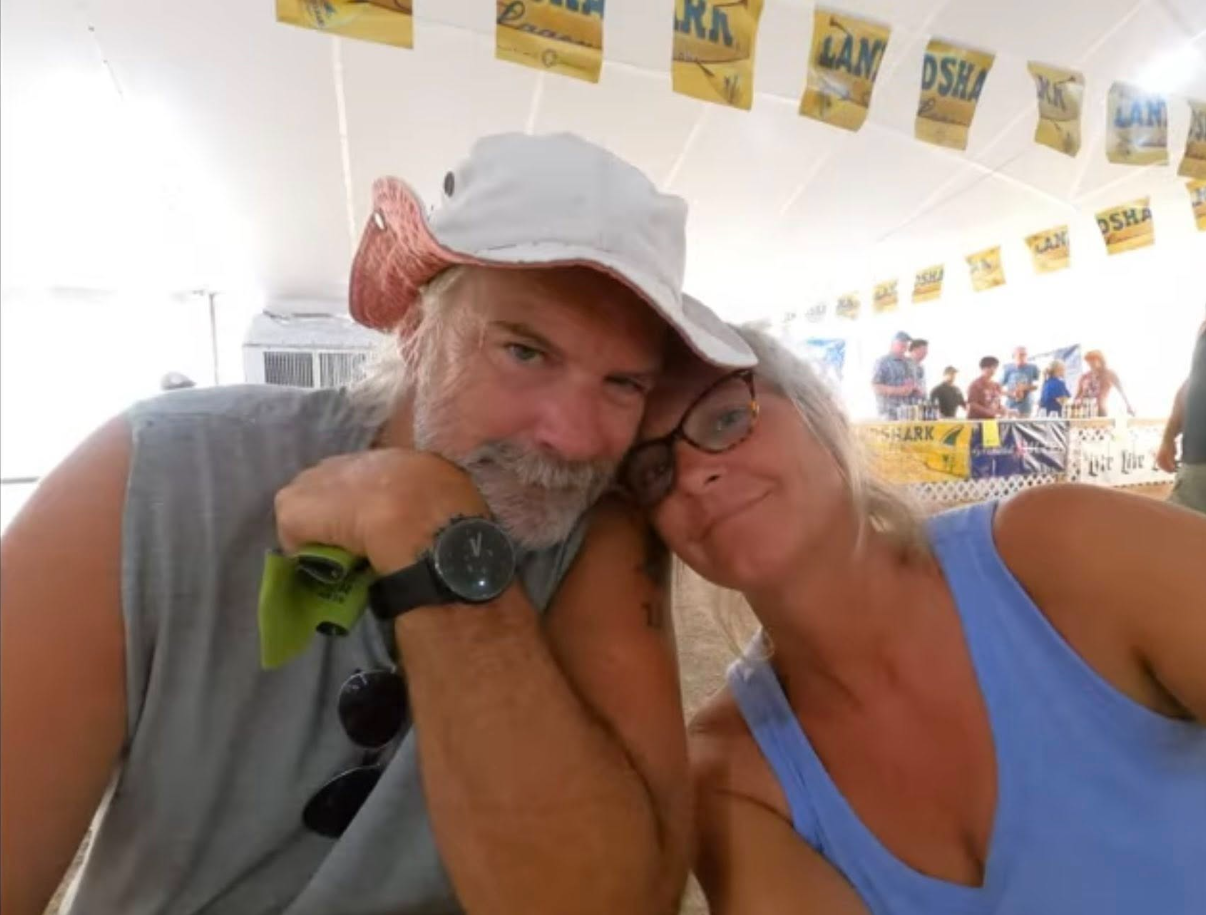 Brian and Lynette Hooker, sit closely together at a casual outdoor event, leaning their heads against each other. | Source: Facebook/PlunderStudios