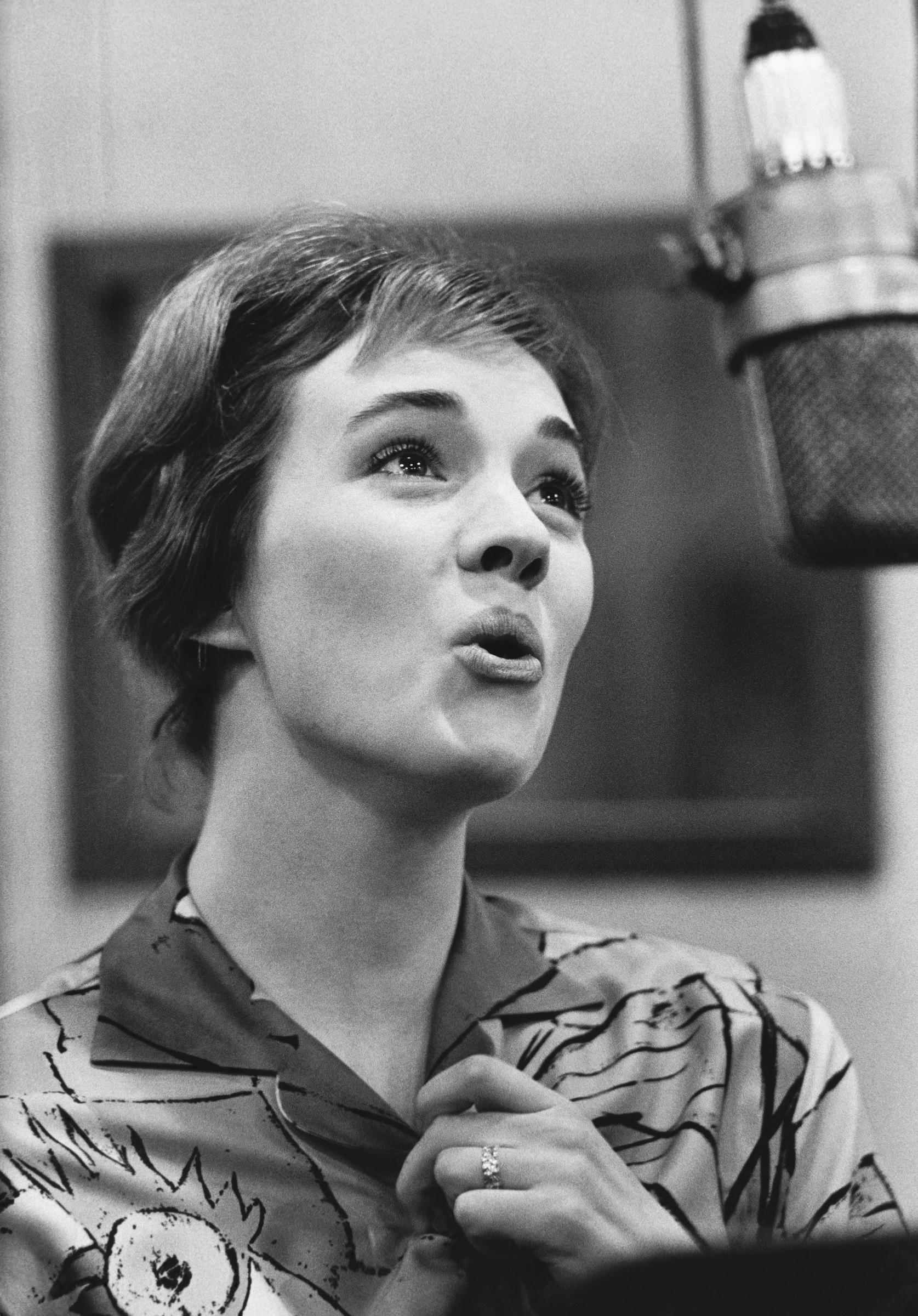 Julie Andrews sings passionately into a studio microphone during a 1961 recording session at the Columbia Records Studio in New York City. The celebrated vocalist wears a collared, abstract-patterned blouse and a sparkling ring, her short, neatly coiffed hair framing an expressive performance behind the mic.