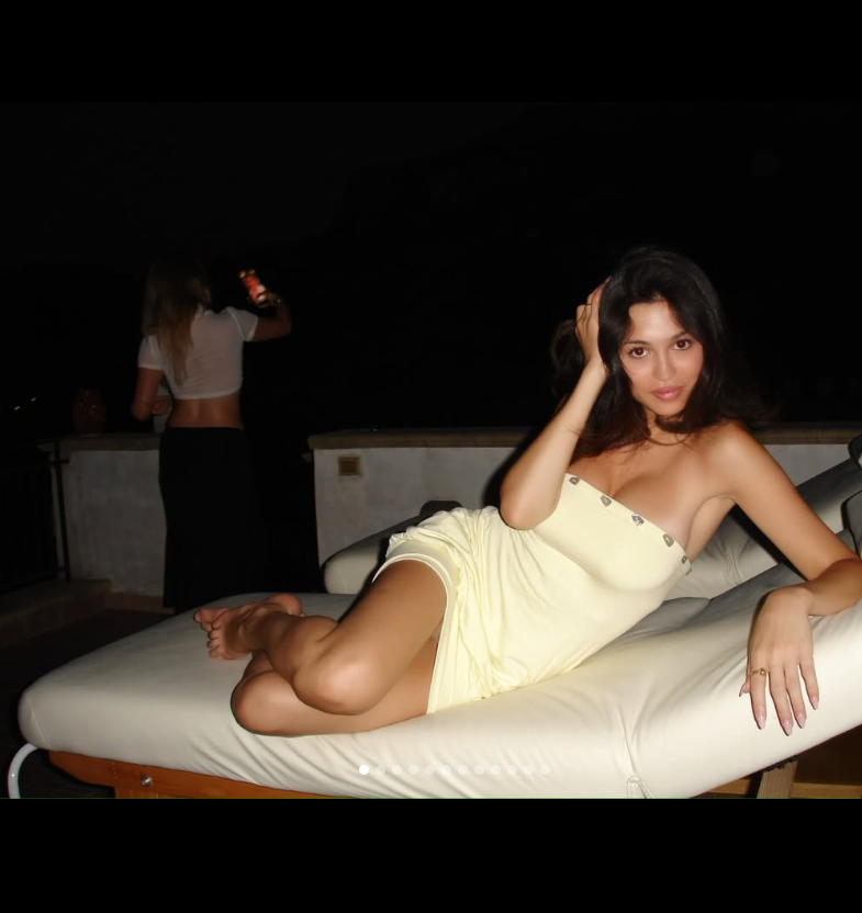 Josie Totah poses on a cushioned outdoor lounge chair at night, wearing a pale-yellow strapless mini dress with decorative buttons along the neckline. She reclines on her side with one hand resting against her head and the other stretched along the chair, while another person stands in the background. | Source: Instagram/josietotah