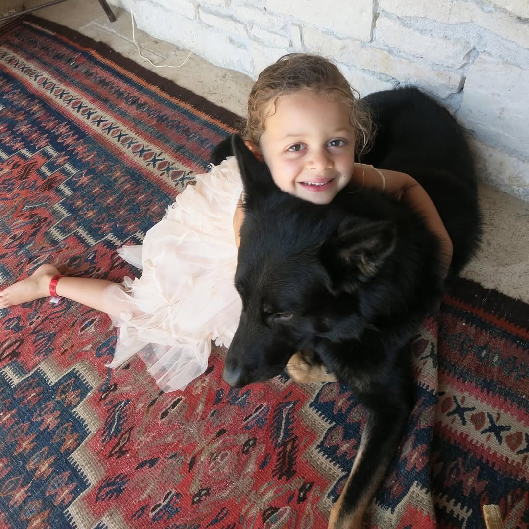 Harlow Winter Kate Madden smiles while sitting on a patterned rug with a large black dog curled against her. Wearing a pale tulle dress, Kate appears relaxed and cheerful in the candid childhood photo.| Source: Instagram/nicolerichie and joelmadden