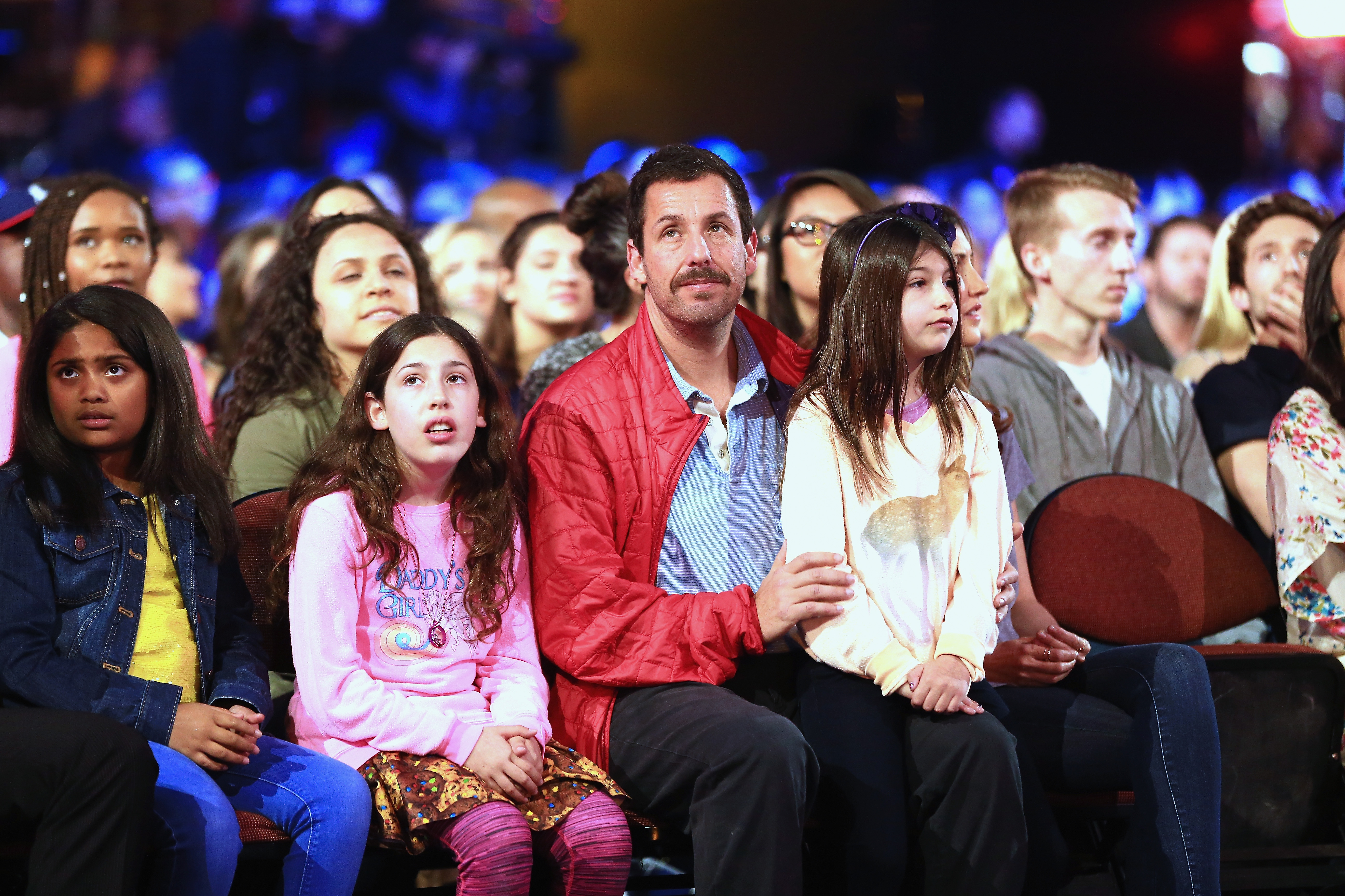 Sadie Madison Sandler sits beside her father Adam Sandler, while Sunny Madeline Sandler leans close as they watch the stage at the Kids’ Choice Awards. Adam, in a red jacket, keeps an arm around Sunny, as both girls look ahead with quiet focus amid the brightly lit crowd.