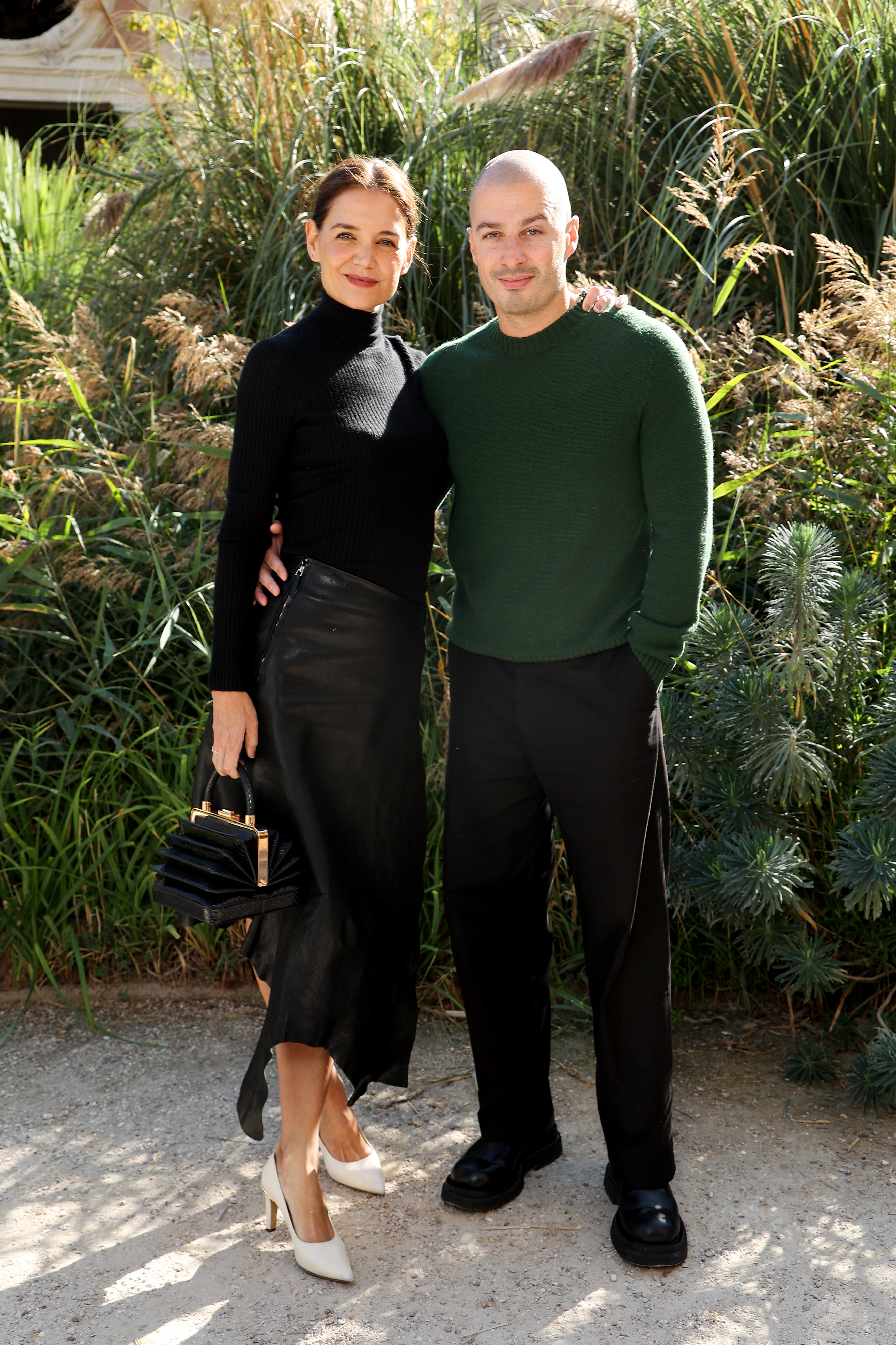 White pointed pumps break up Katie Holmes' otherwise black outfit, while Jason Bard Yarmosky's forest-green sweater adds the strongest color beside her. The actress and artist arrived together for Gabriela Hearst's Spring/Summer 2027 show in Paris.