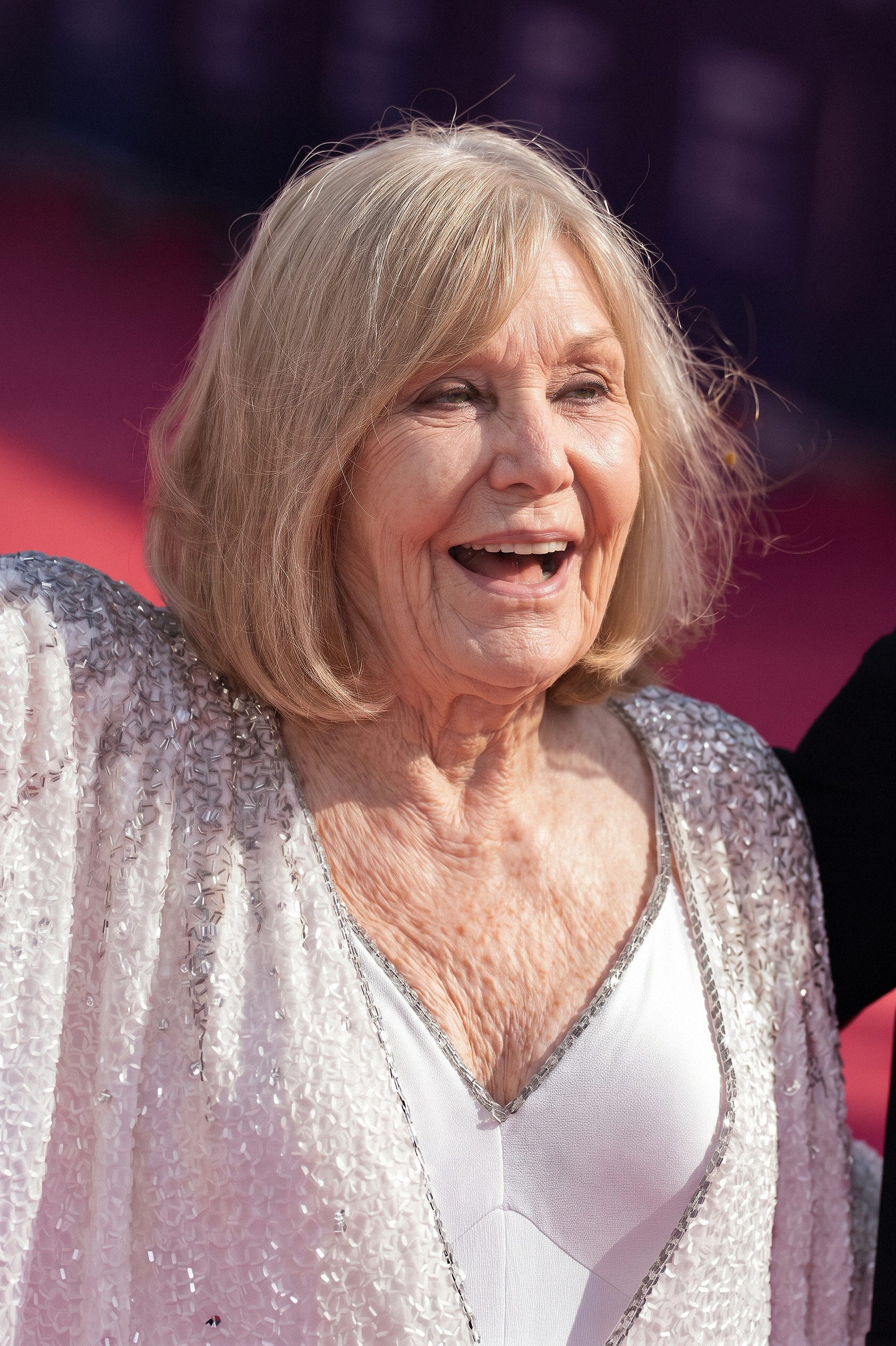 Kim Novak smiles broadly in a sparkling silver jacket over a white V-neck dress, with her blonde bob framing her face against a pink-and-purple event backdrop. | Source: Gemini