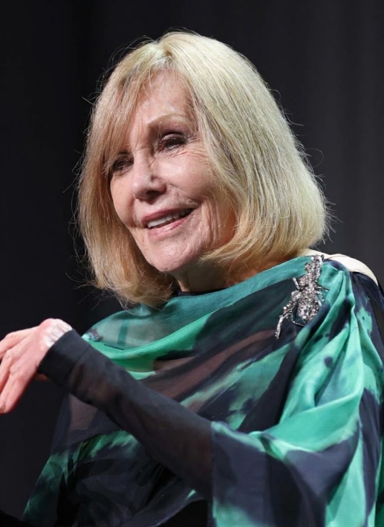 Kim Novak smiles during a stage appearance in a flowing green-and-black outfit, with sheer sleeves and a sparkling brooch adding detail to her look. | Source: Gemini