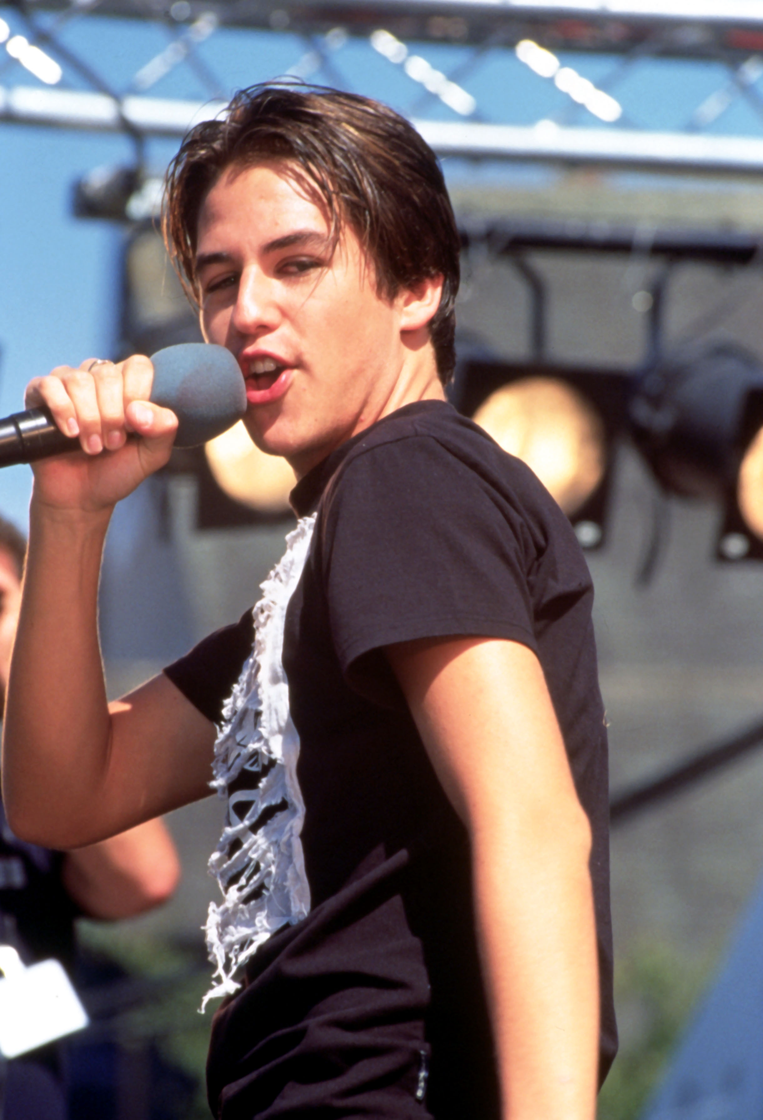 Still in his teens in 1997, he commands the stage in a live performance, wearing a black T-shirt with a white design on the front. With tousled hair and a natural ease, he looks completely in his element.