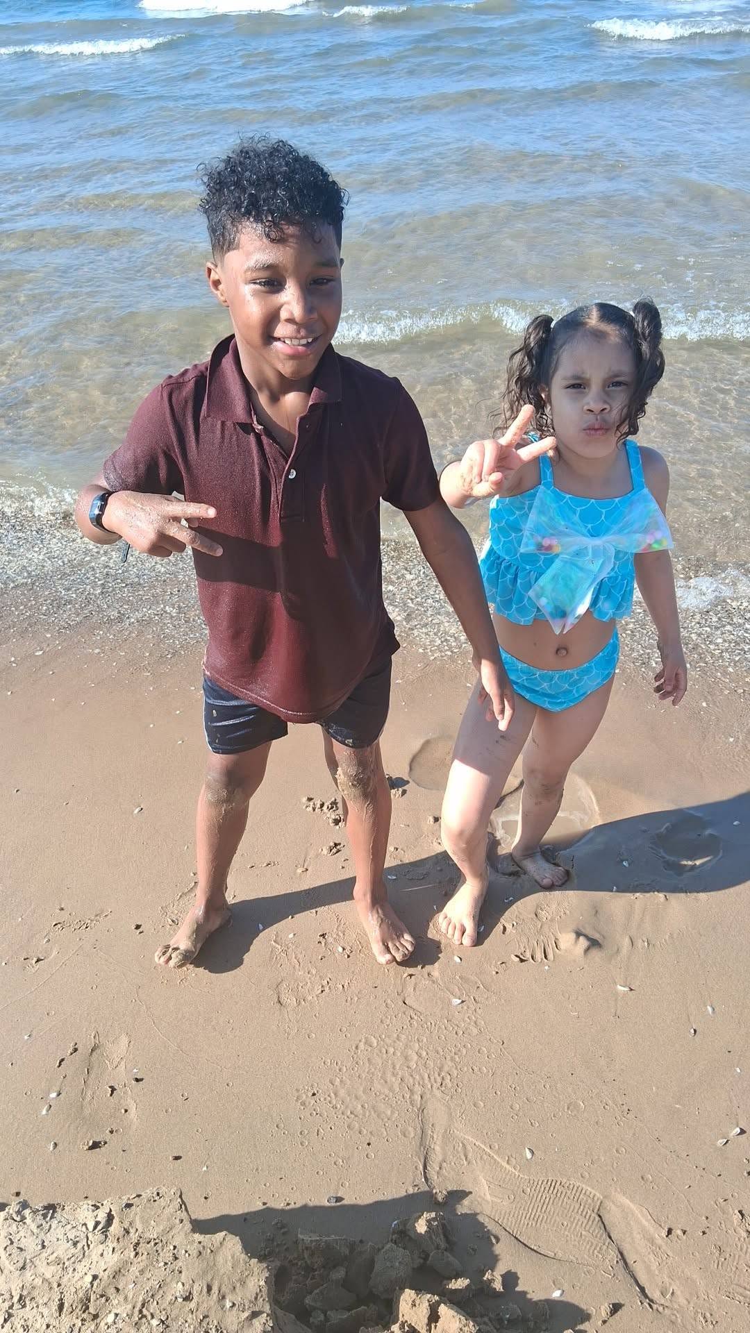 Eddimar Paola Hernández Chacón and her brother stood barefoot along the wet shoreline on a sunny beach outing. Eddimar held up a peace sign while wearing curly pigtails and her turquoise mermaid-scale two-piece swimsuit with an iridescent bow, posing beside her brother, who smiled in a maroon collared polo shirt and dark swim trunks against the gentle ocean waves. | Source: Facebook/Morena Esa
