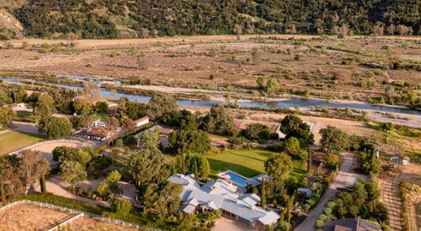 An aerial view of the property reveals its landscaped grounds, neighboring homes, and the river valley stretching toward the surrounding hills. | Source: Redfin