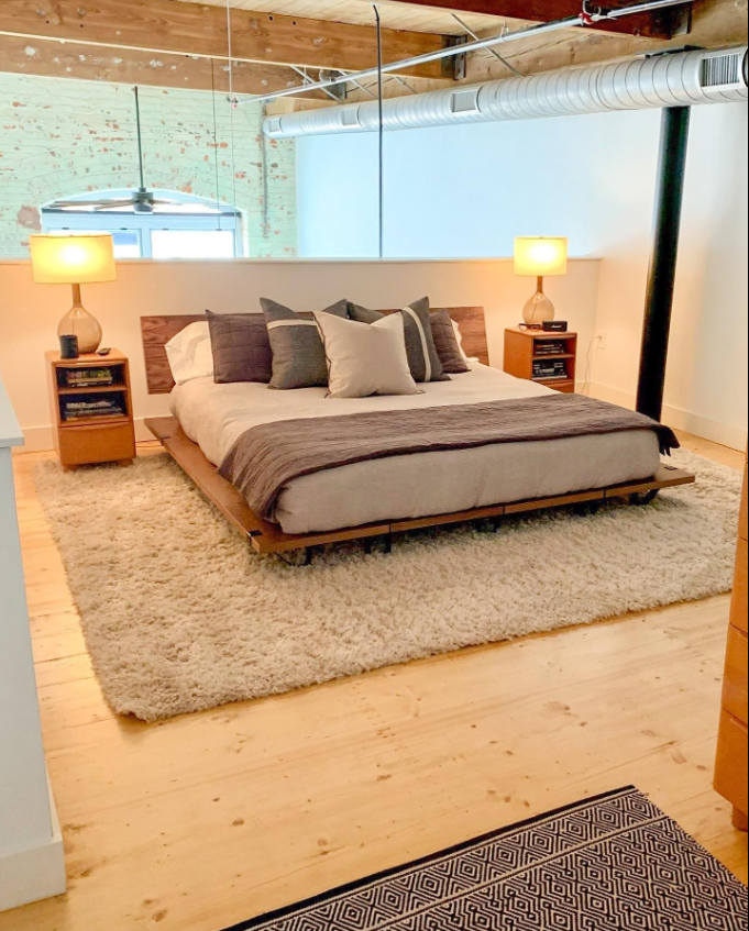 A warm bedroom setup pairs a low wooden platform bed with layered gray-and-white bedding, matching nightstands and a plush cream rug beneath it. | Source: Facebook/Judson Mills Loft