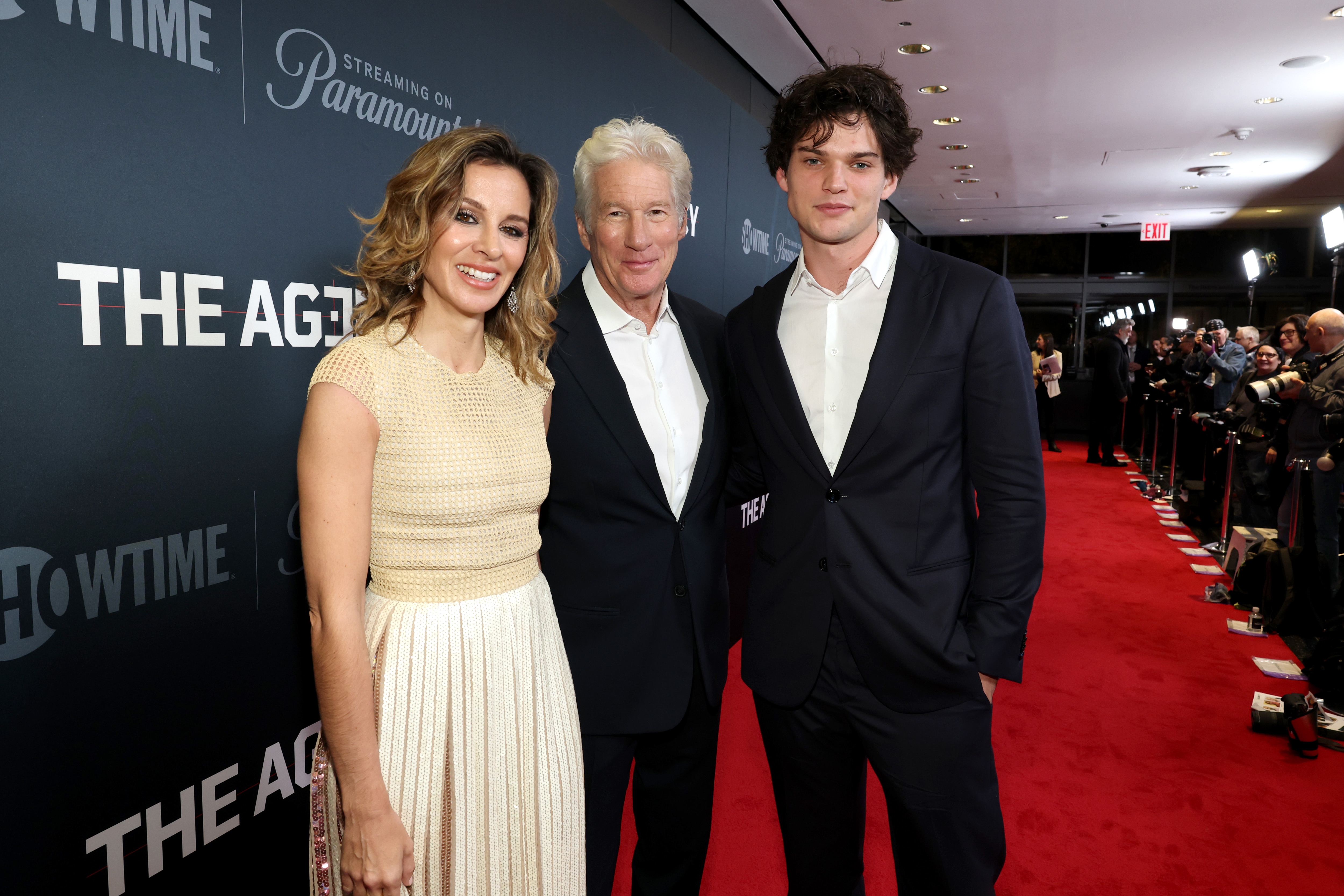 All smiles and sophistication, Homer Gere stands between Alejandra and Richard Gere at The Agency premiere. Cameras flash as the trio lights up the red carpet in New York City.