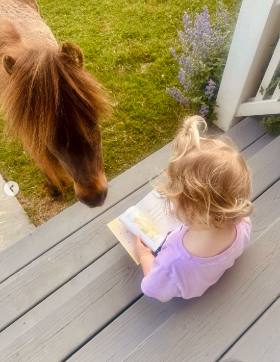 Kaliyah sits on wooden porch boards with an open picture book in her lap as a small brown pony stands beside her. The quiet scene offers a contrast to the tractor, horseback rides, and backstage photographs. | Source: Instagram/wynonnajudd