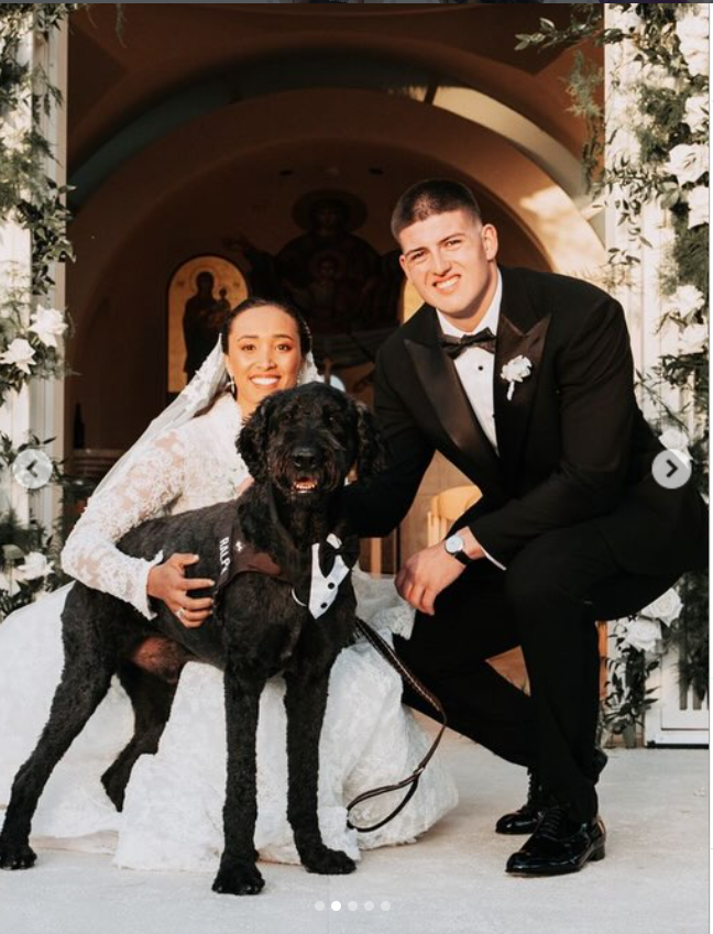 George Karlaftis and Kaia Harris pose outside a church entrance on their wedding day alongside their black dog, who wears a tuxedo-style harness. | Source: Instagram/georgekarlaftis