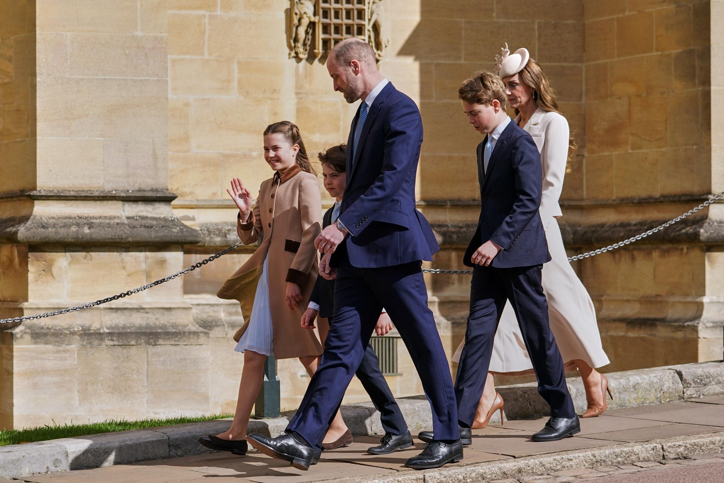 Shot in profile along the sun-drenched exterior of St George's Chapel, this image shows the Wales family in full stride, the Prince of Wales leading the group with Prince Louis tucked close beside him. Princess Charlotte waves cheerfully to onlookers on the left, her blue pleated dress lifting in the breeze beneath her camel coat, while Prince George and the Princess of Wales follow just behind. | Source: Getty Images