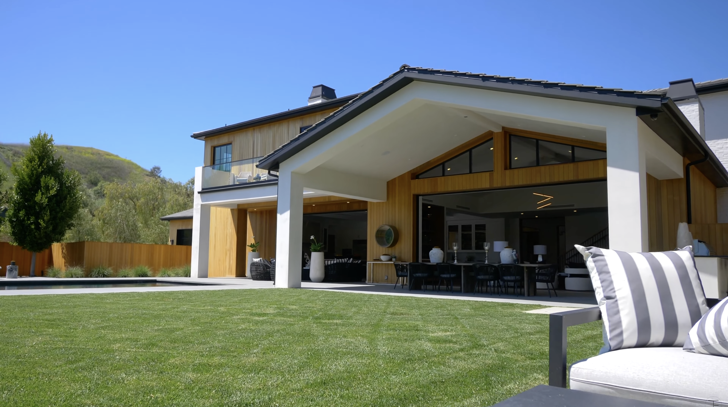 The rear of the house opens onto a large lawn, with a deep covered patio providing space for outdoor dining. Wood siding and black-framed openings add contrast to the white exterior. | Source: Youtube/Tomer.Fridman
