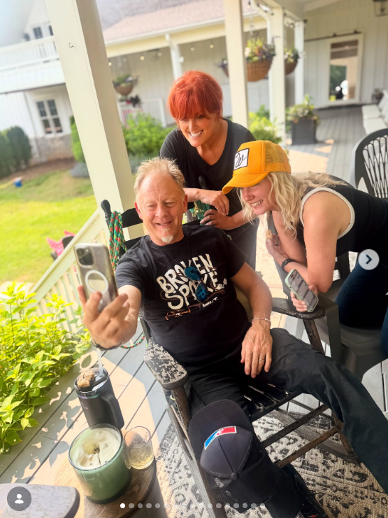 Wynonna leans toward Cactus Moser as he holds up a phone for a photograph with another woman. Hanging plants, outdoor seating, and the lawn beyond the porch create a relaxed setting for the gathering. | Source: Instagram/wynonnajudd