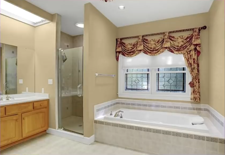 The primary bathroom features a built-in soaking tub beneath a window, a separate glass-enclosed shower, and a wooden vanity with a light countertop. Beige walls, decorative curtains, and tiled surfaces give the room a classic, traditional look.